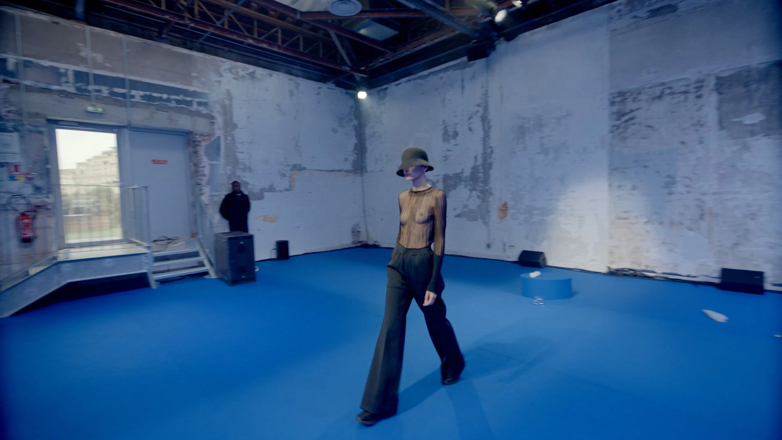 An art installation or mannequin display in a large, industrial-style room with unfinished walls and blue flooring. The mannequin is wearing wide-leg pants, a striped long-sleeve top, and a wide-brimmed hat, with a partly painted or covered face. The