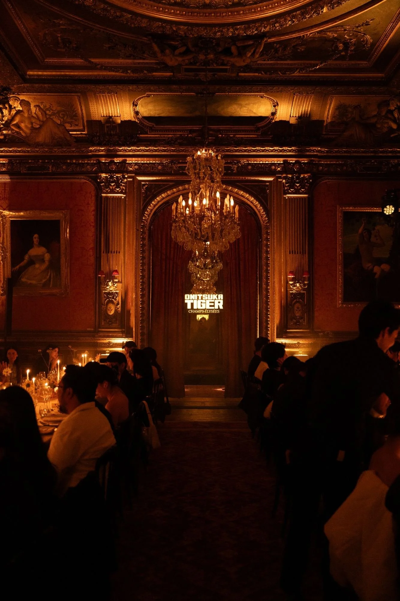Luxurious ballroom with ornate gold detailing, large chandelier, and portraits on the walls, filled with elegantly dressed people sitting at tables, softly lit with candles.