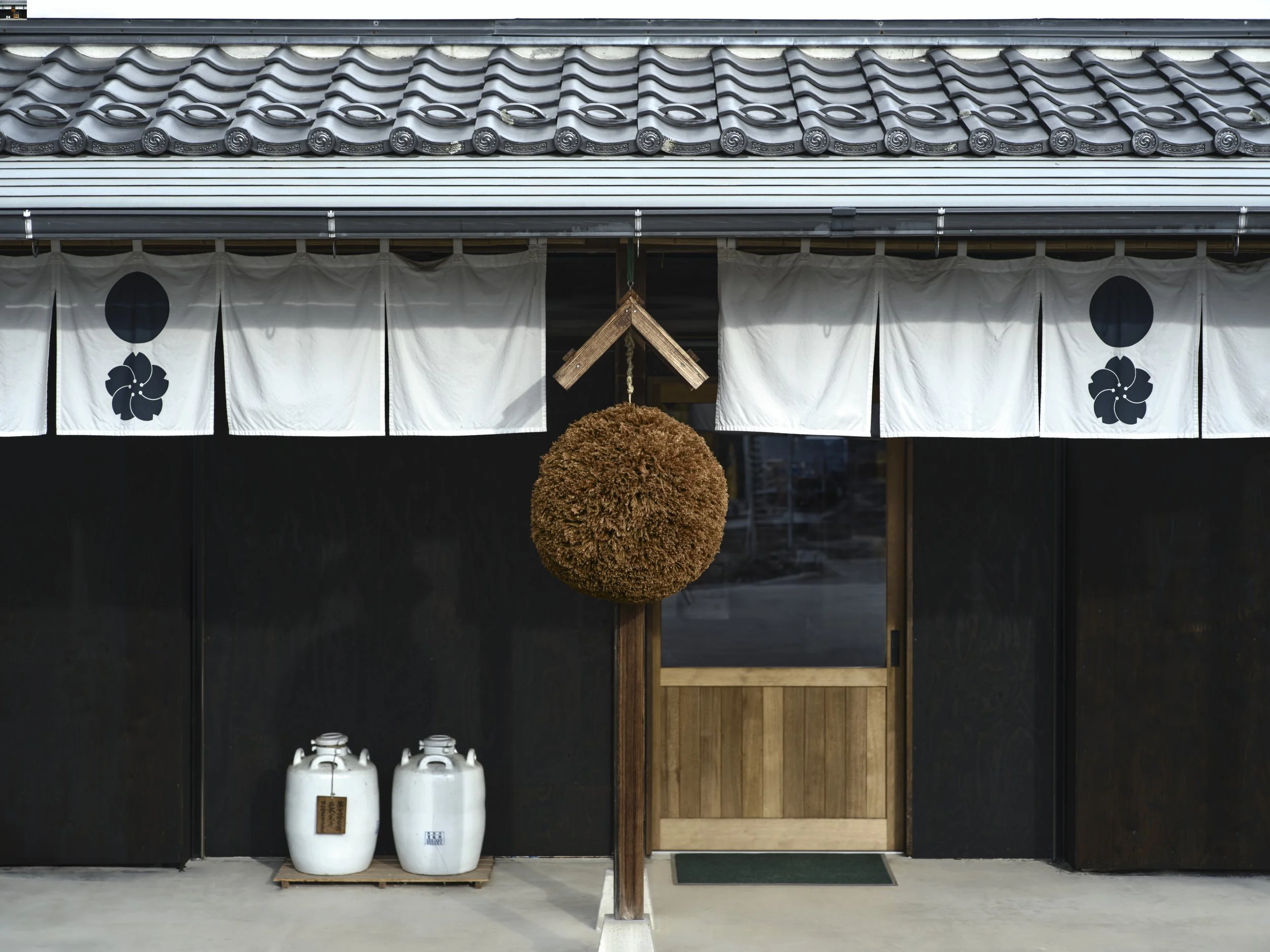 Traditional Japanese entrance with white noren curtains featuring black symbols, a large ornamental ball, and a wooden structure. Propane tanks are visible at the bottom left.