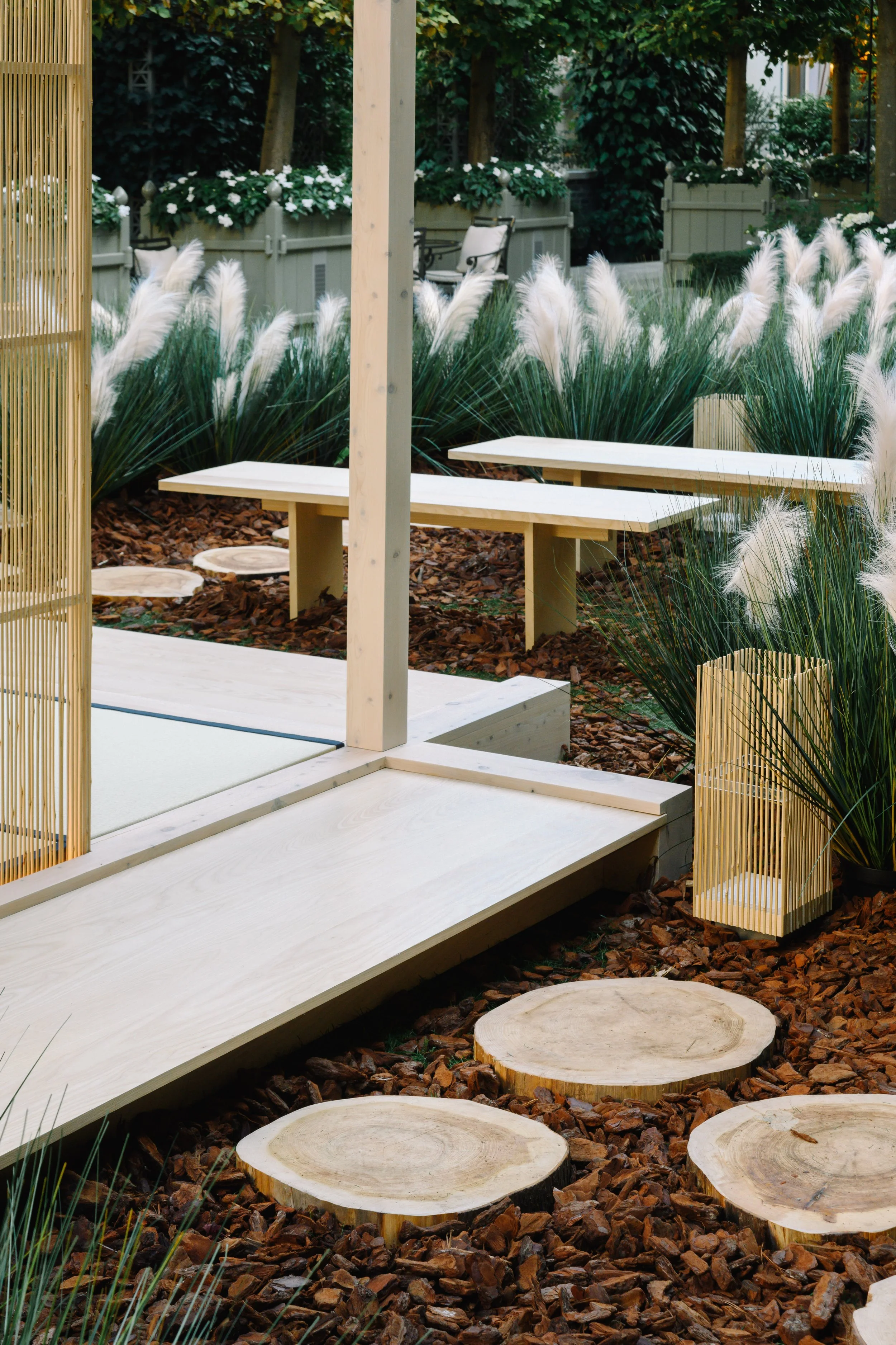 Garden scene with white bench, round stone stepping stones, ornamental grass, and wooden fencing and structures.