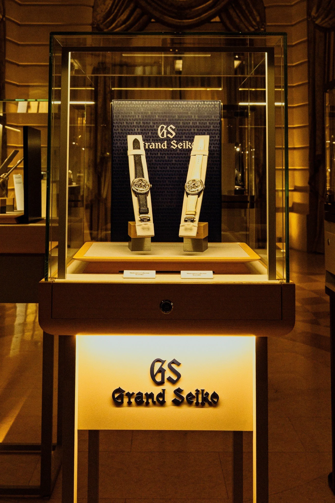 Display case featuring two Grand Seiko watches, one with a black strap and the other with a gold strap, inside a luxury retail store.