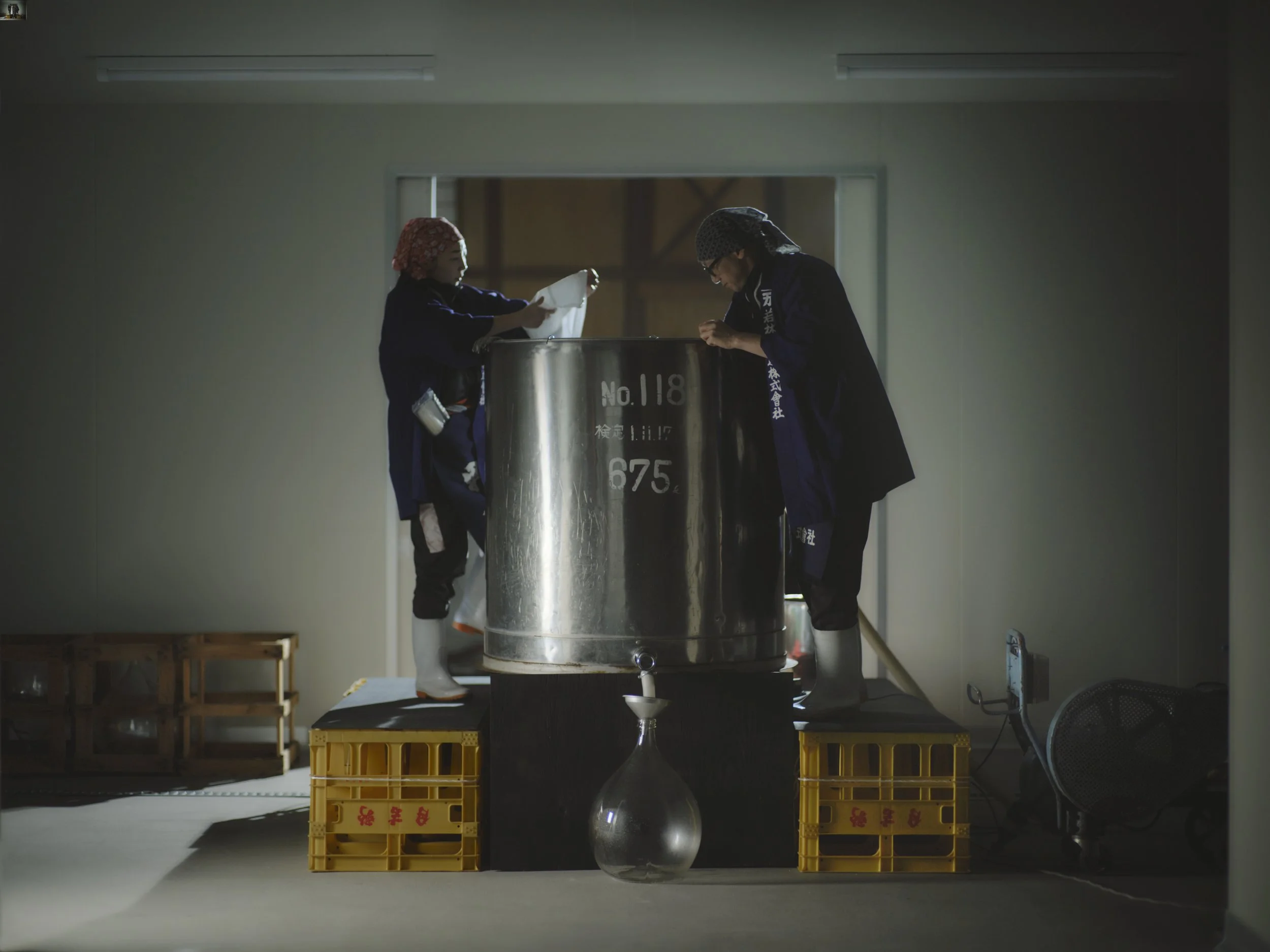 Two people wearing hair coverings and blue coats are working with a large metal container while standing on yellow crates in a room with plain walls and a window. Sunlight illuminates the scene from behind.