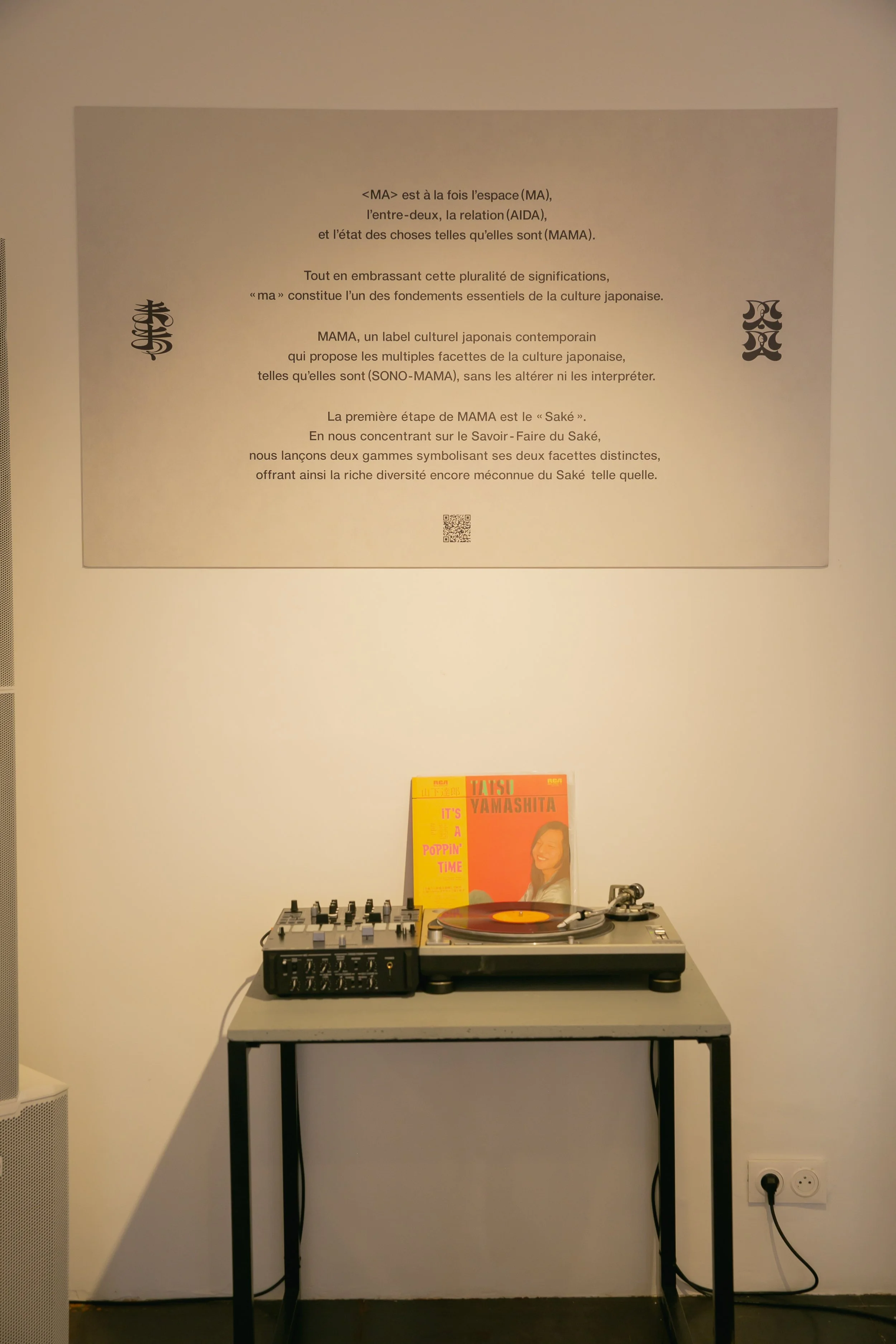 A DJ turntable setup on a small black table with a mix board to the left, a vinyl record playing, and a yellow and orange album cover behind it. A large informational poster with French text about the cultural significance of 'Ma' and 'Saké' hangs on