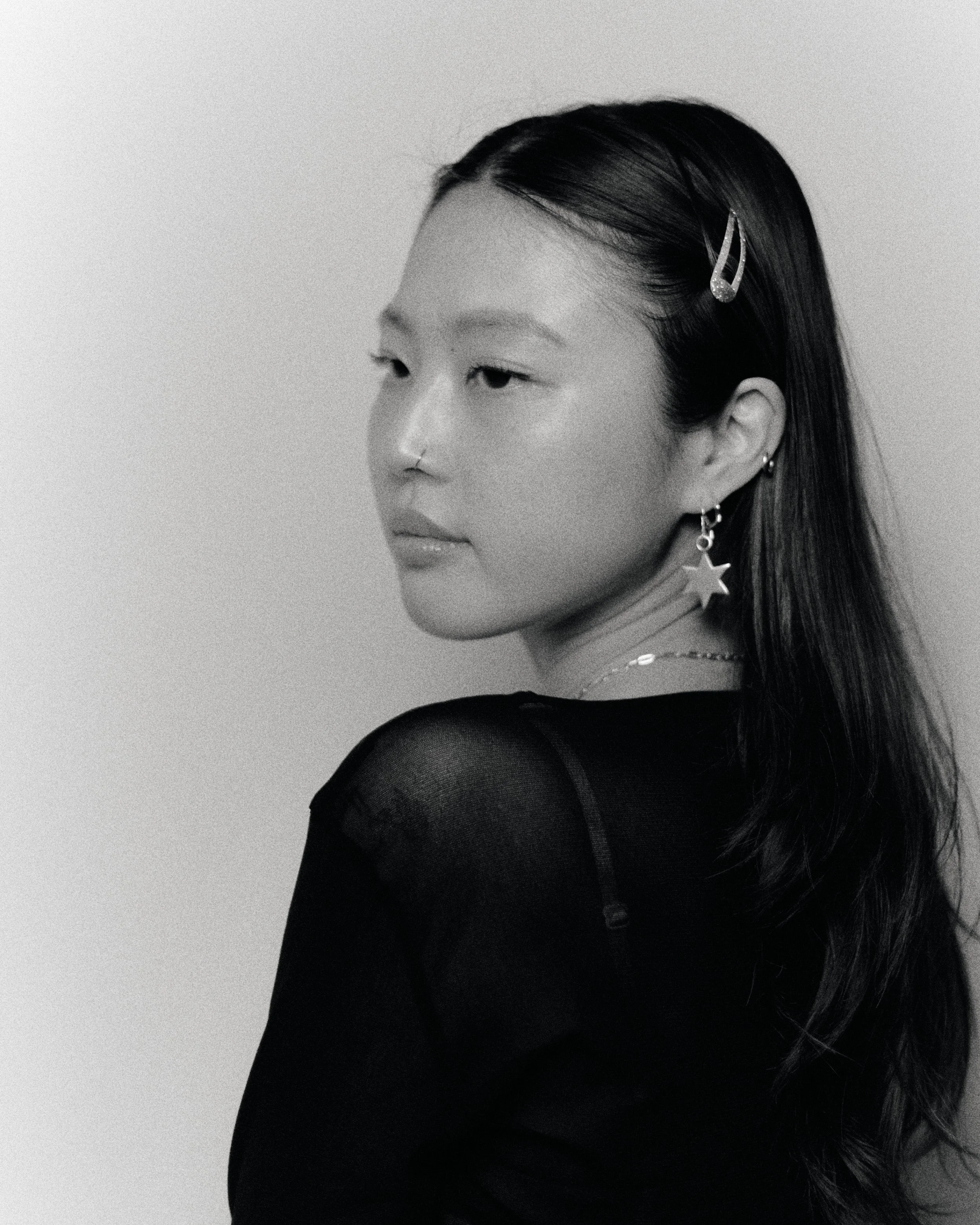 Black and white portrait of a woman with long dark hair, wearing star-shaped earrings, a hair clip, and a high-neck top, posed against a light-colored background.