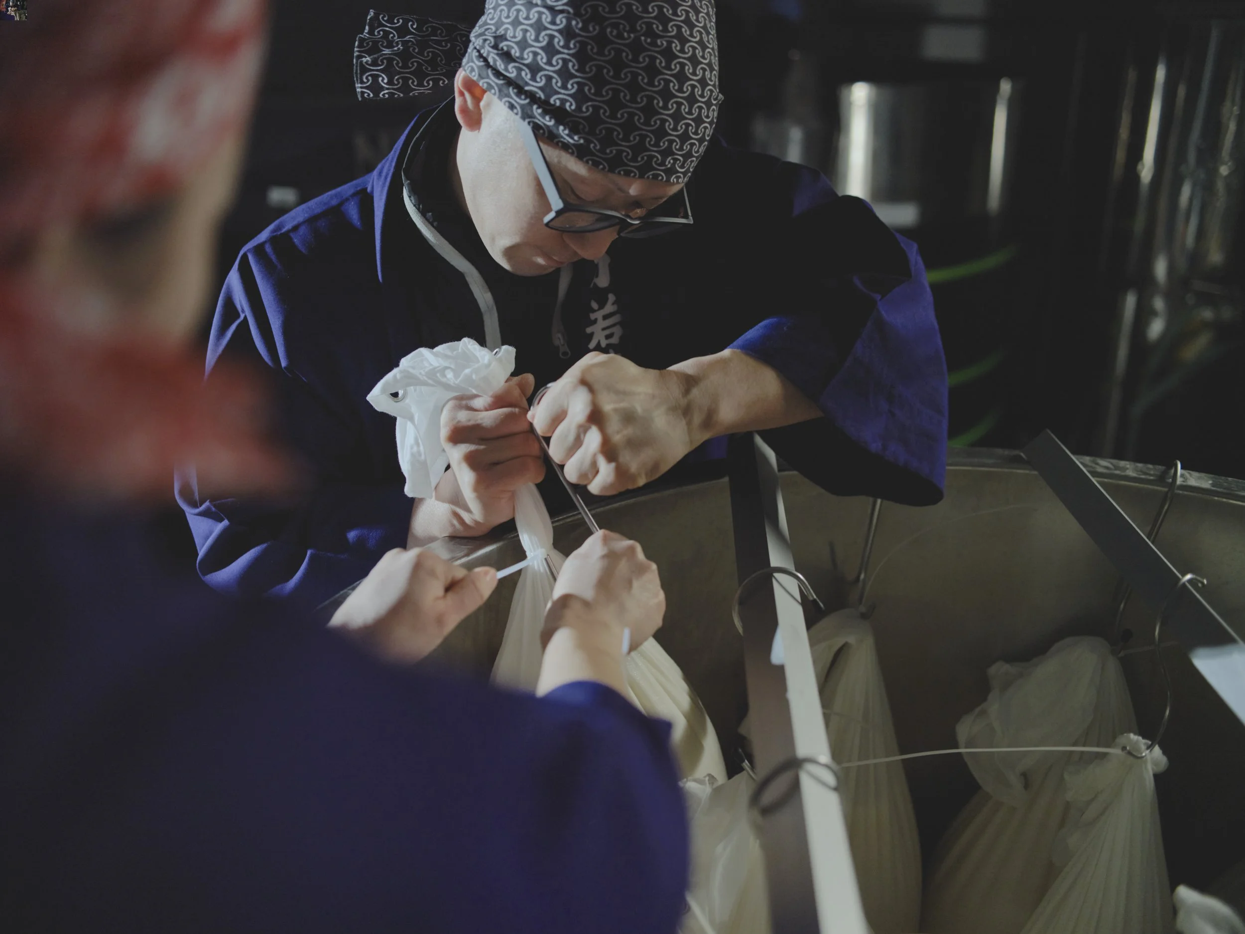 A chef wearing glasses, a bandana, and a blue jacket works carefully on a culinary project.