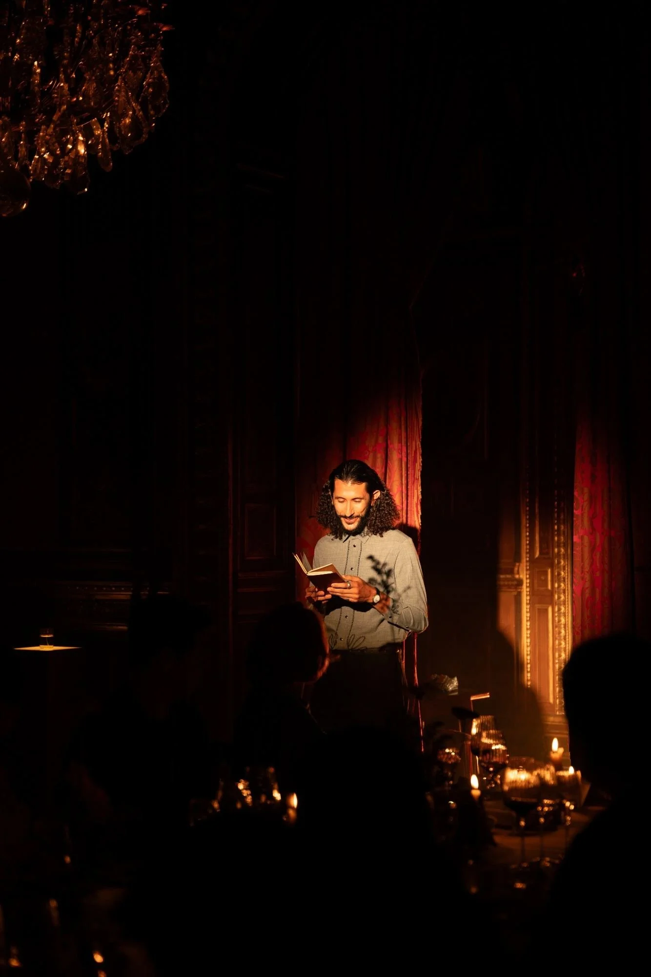 A man with long curly hair standing on a dimly lit stage, reading a book, with candles on a table in the foreground and a chandelier hanging from the ceiling in the background.