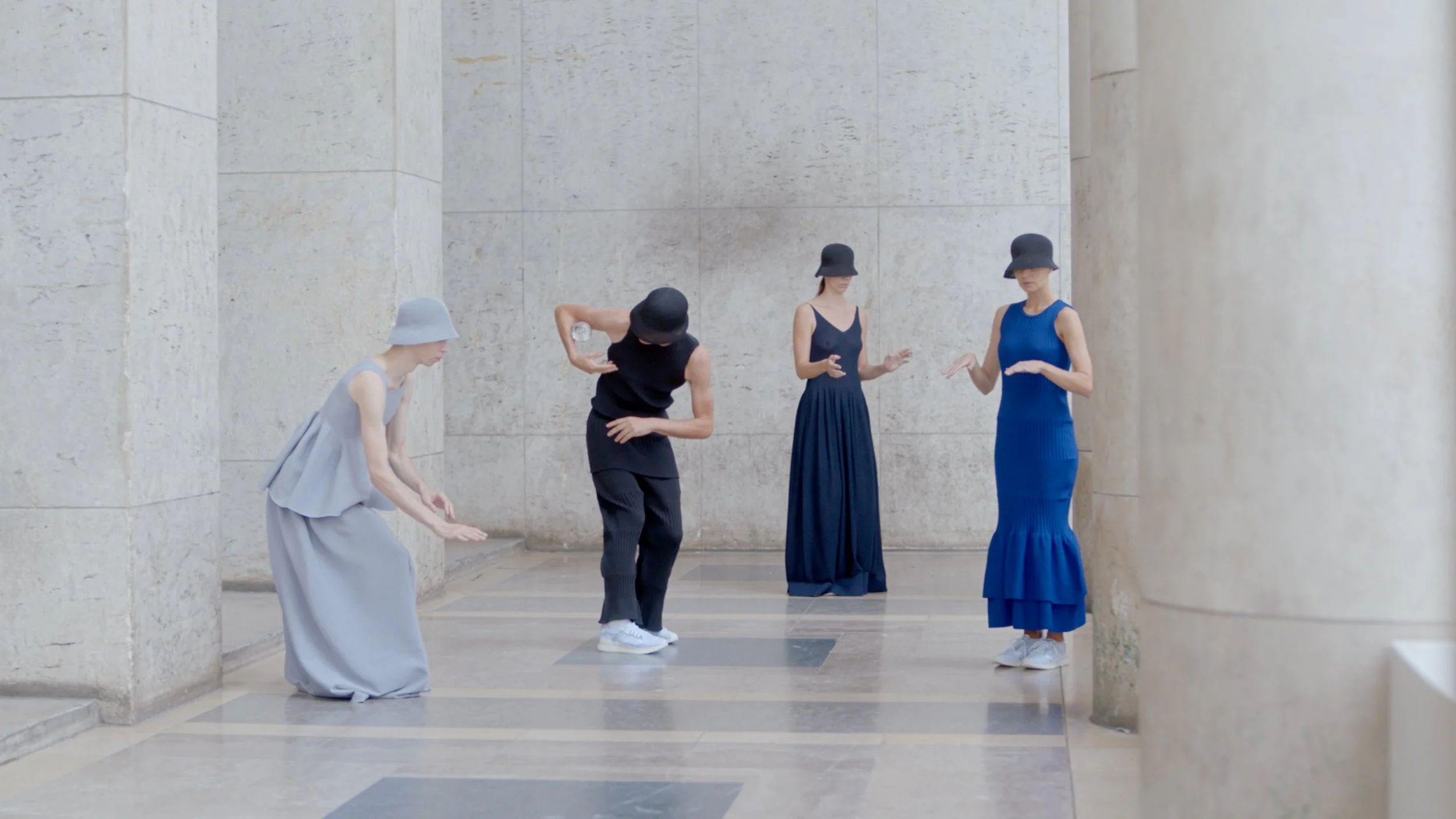 Five women wearing elegant dresses and black hats, standing and leaning against a stone wall with columns, in an outdoor or semi-outdoor setting.
