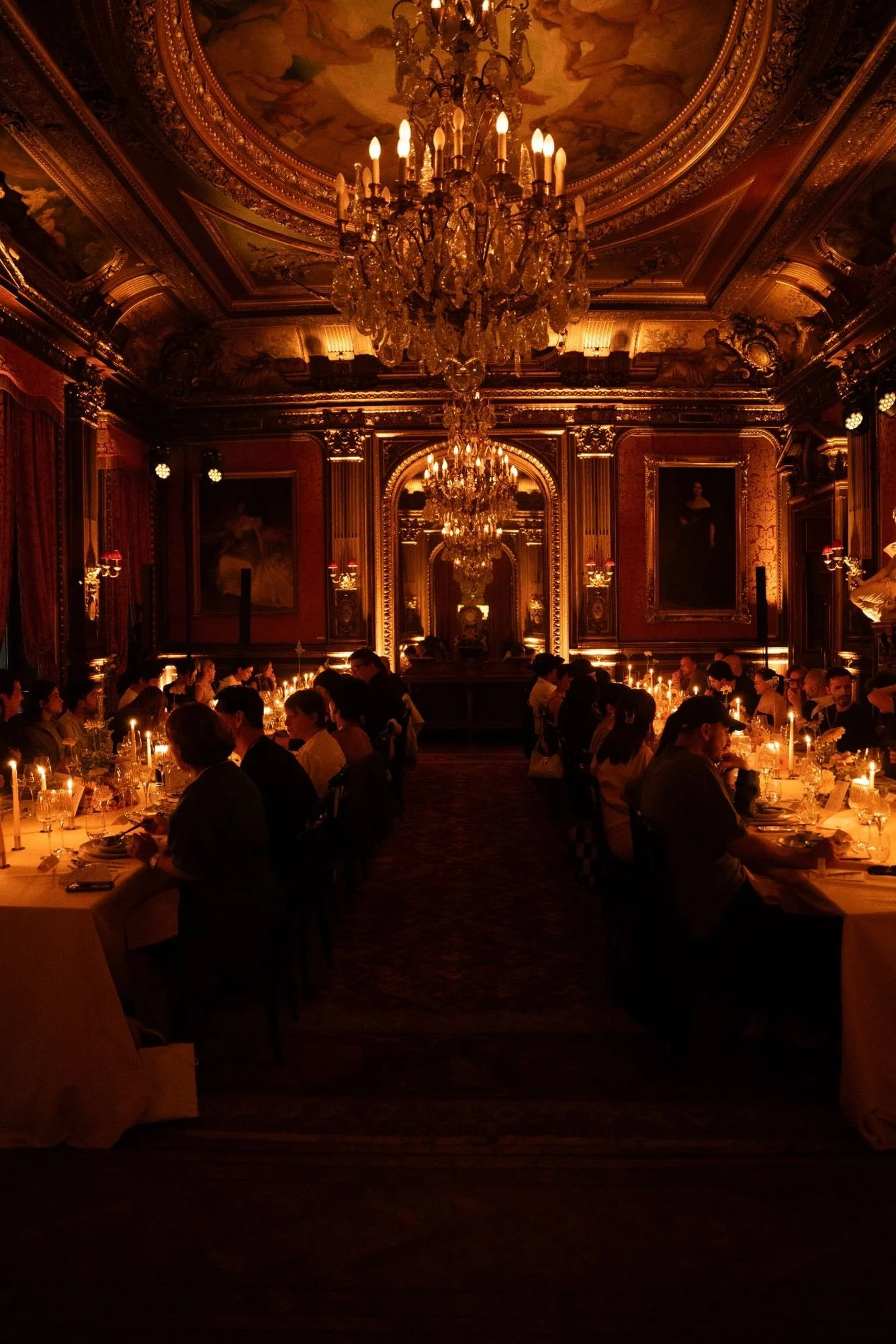 Elegant dining room filled with guests, ornate gold decorations, hanging chandeliers, and dim candlelight.