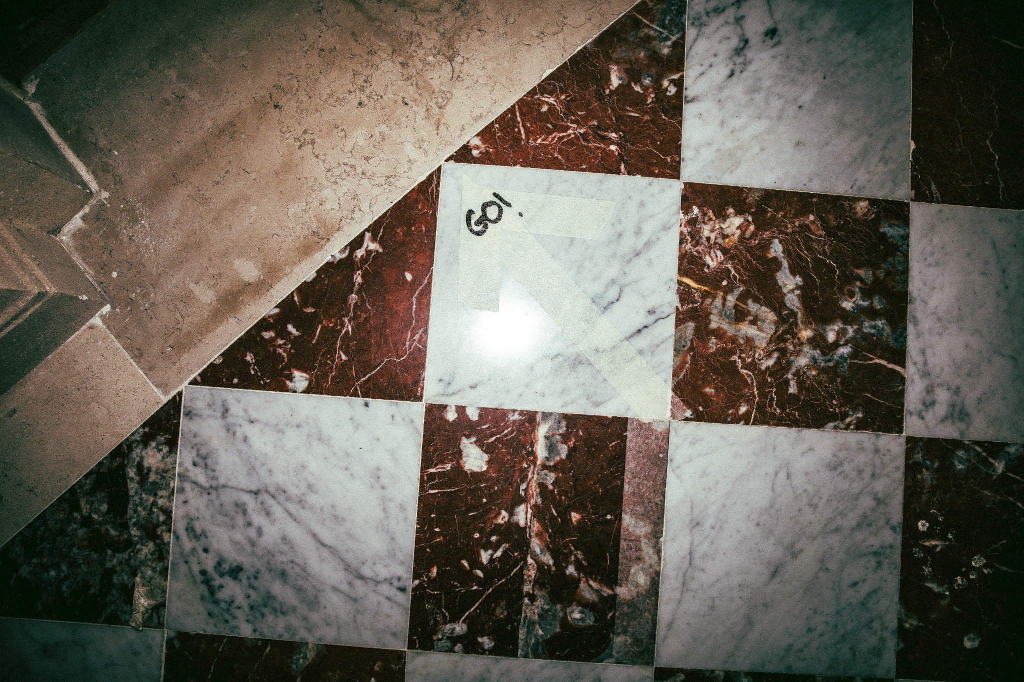 A close-up of a floor with square marble tiles in various colors and patterns, with a piece of tape labeled 'GO!' on one tile.