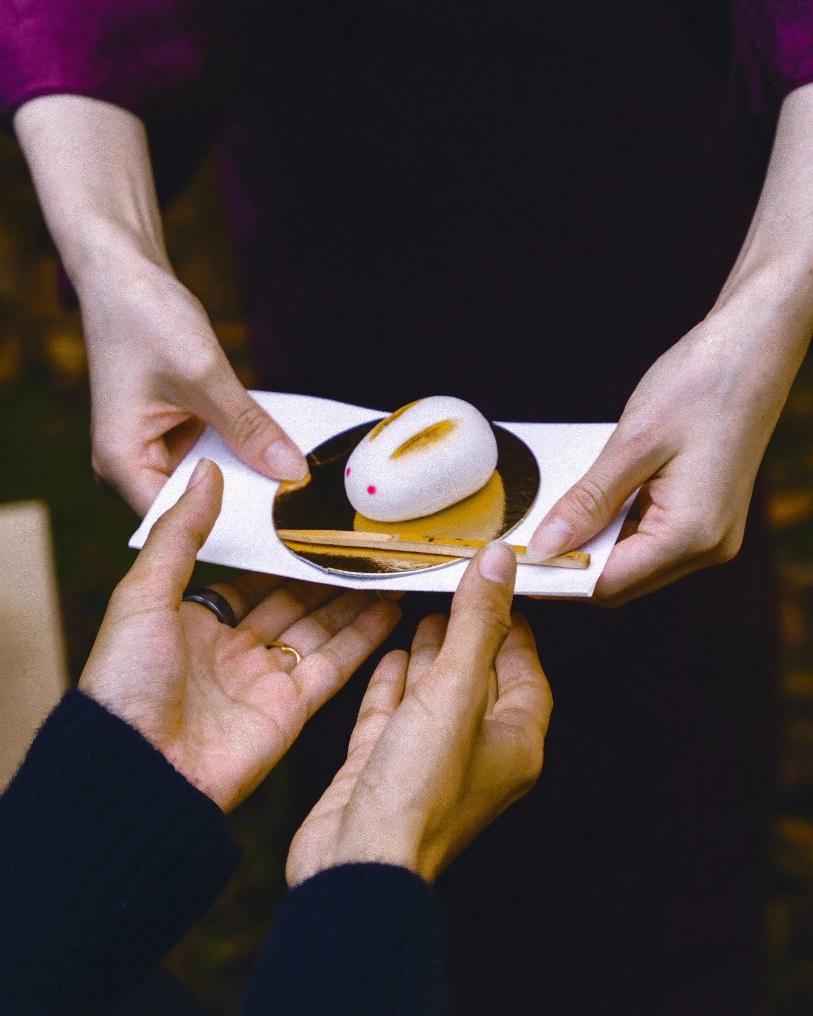 Close-up of hands wrapping a dessert, possibly a mochi, on a dark plate with a person in a black and purple outfit in the background.