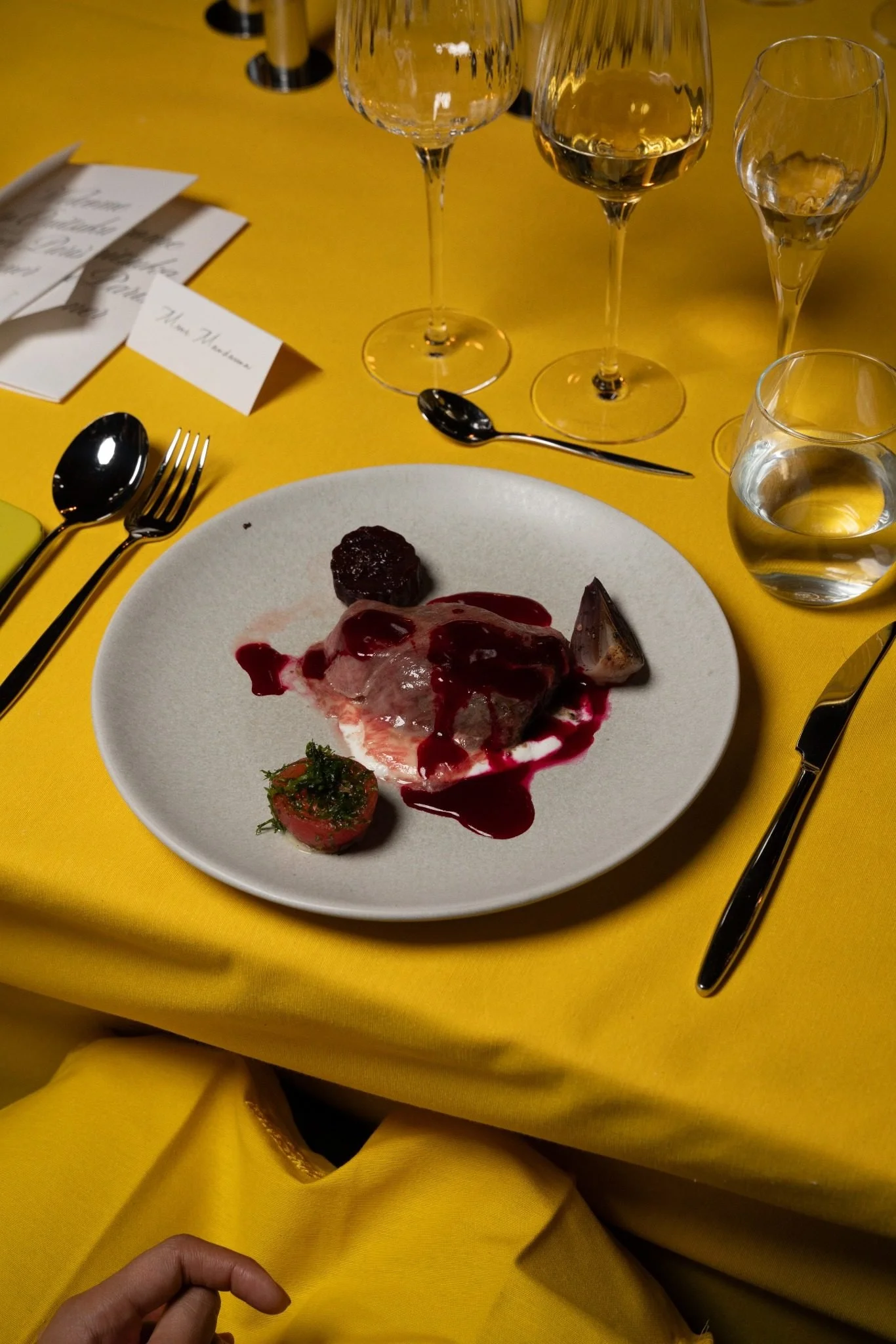 A dining table with a white plate holding a cooked meat dish with red sauce, a small roasted vegetable, and greens. There are wine glasses, a water glass, utensils, and place cards on a yellow tablecloth.