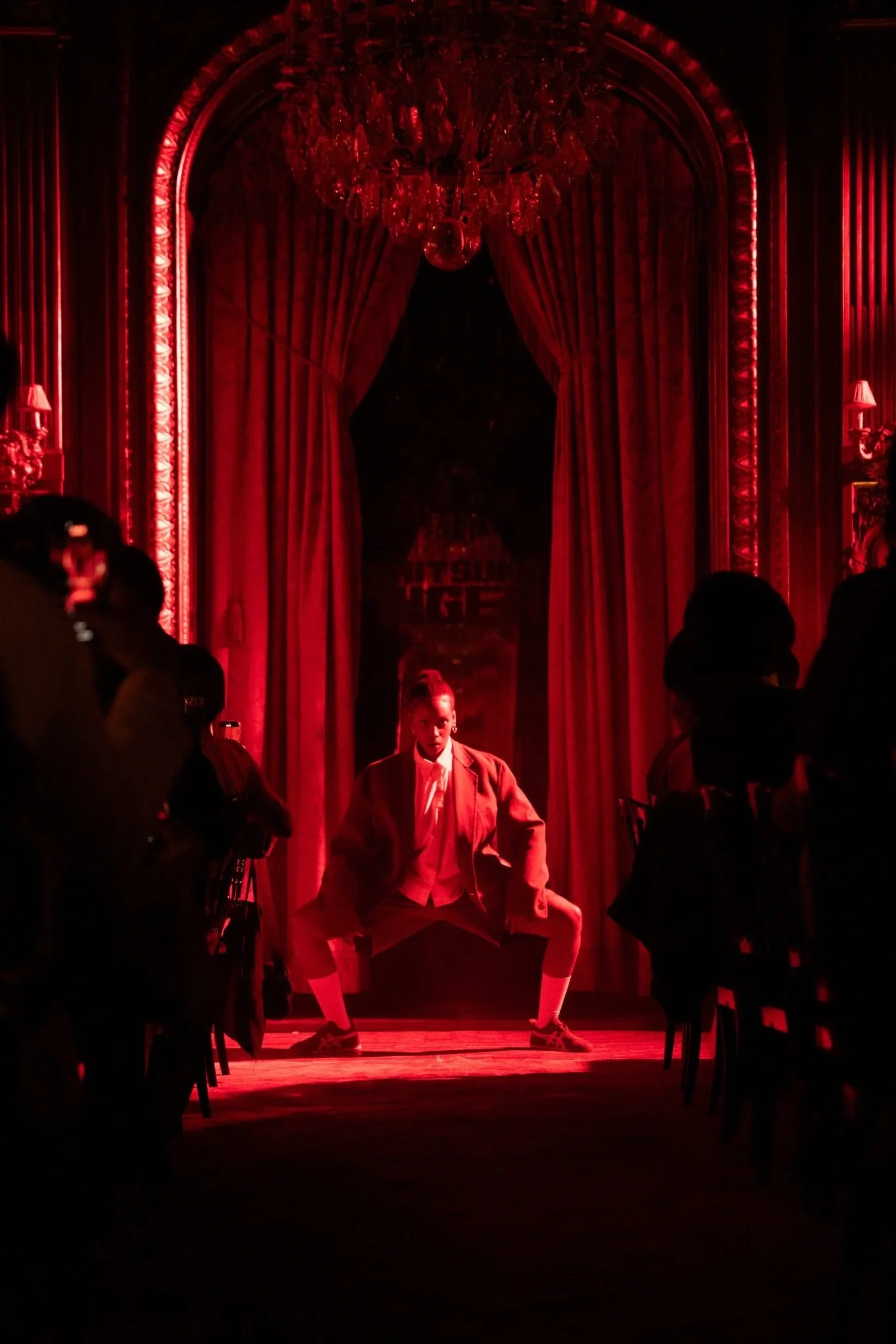 A performer in a suit poses on stage under red lighting in a theater with ornate red curtains and chandeliers, with audience members visible in the foreground.