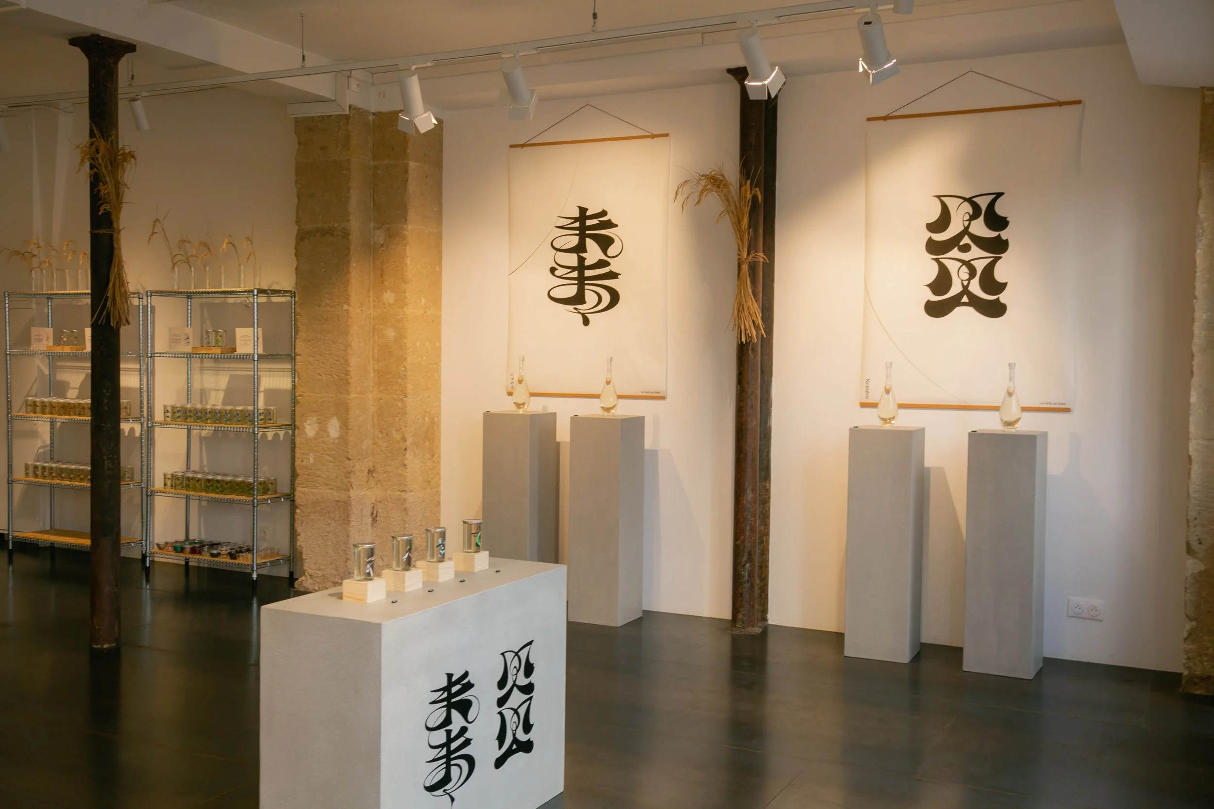 Art exhibit with three vertical banners featuring black Asian script on a white background, sculptures on pedestals, and a shelving unit with small items.