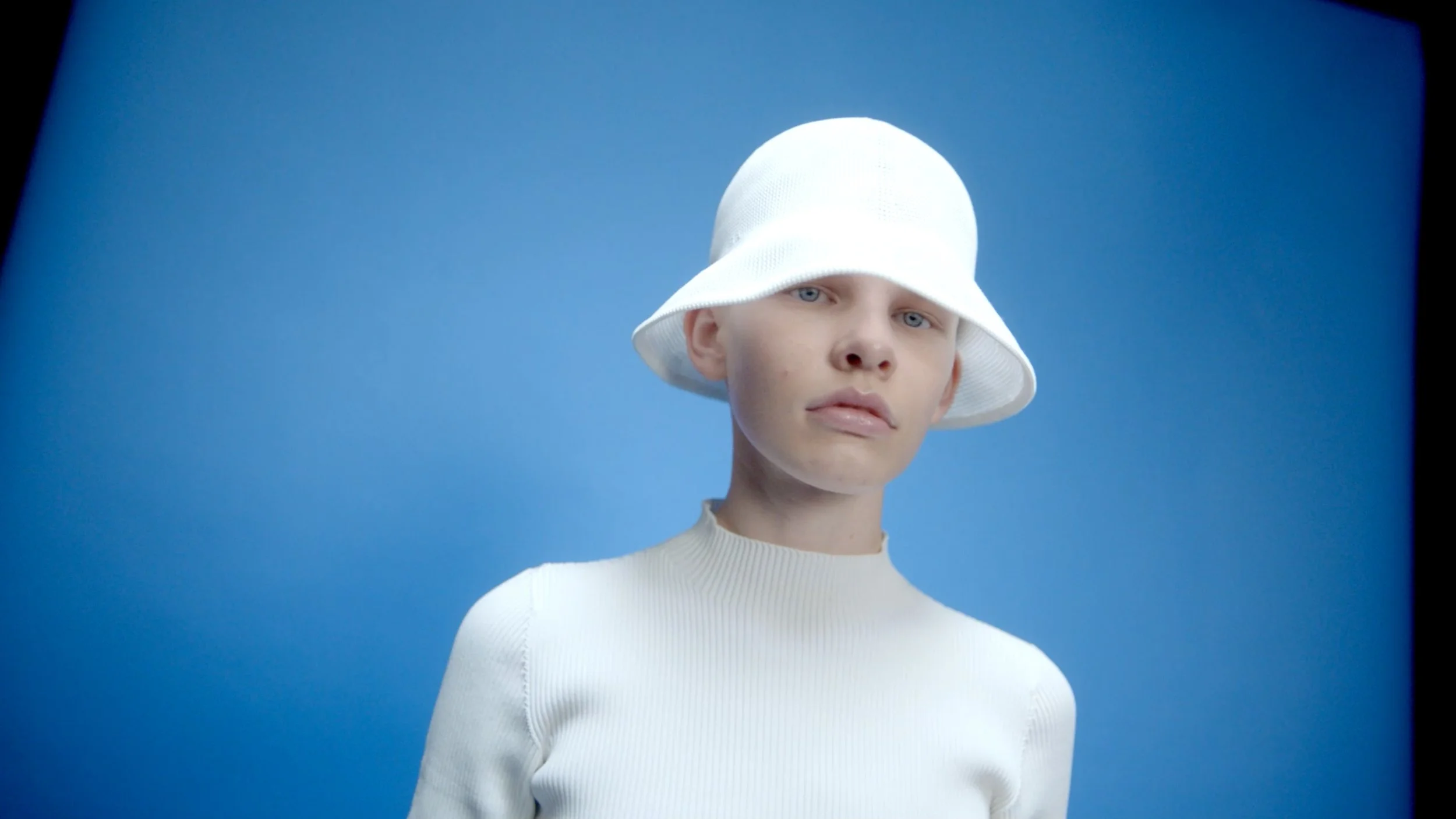 A person with light skin and blue eyes wearing a white hat and white sweater against a blue background.