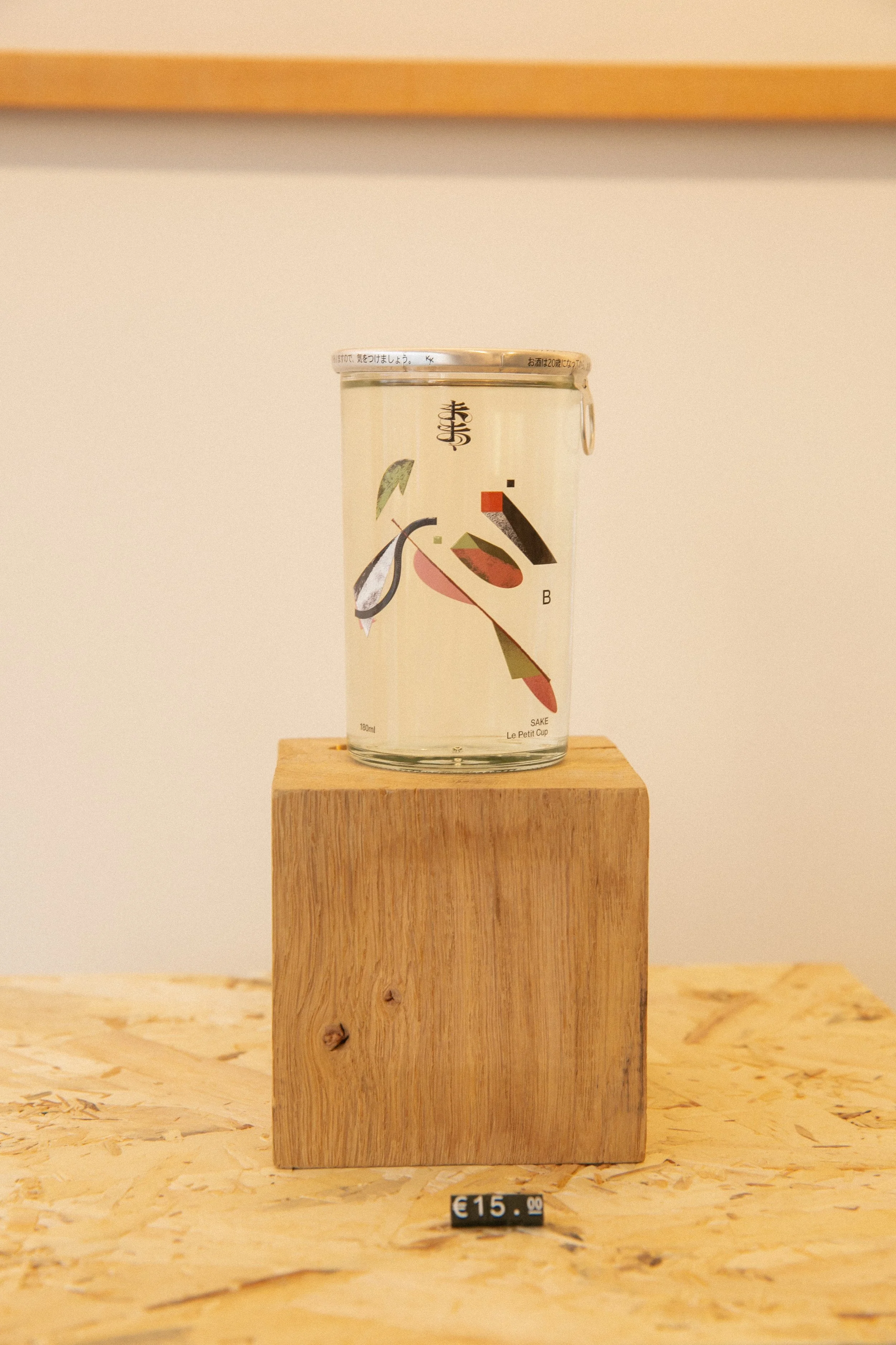 A can of Japanese sake sitting on a wooden block, with a small price tag of 15 euros in front of it, in a minimalist setting.