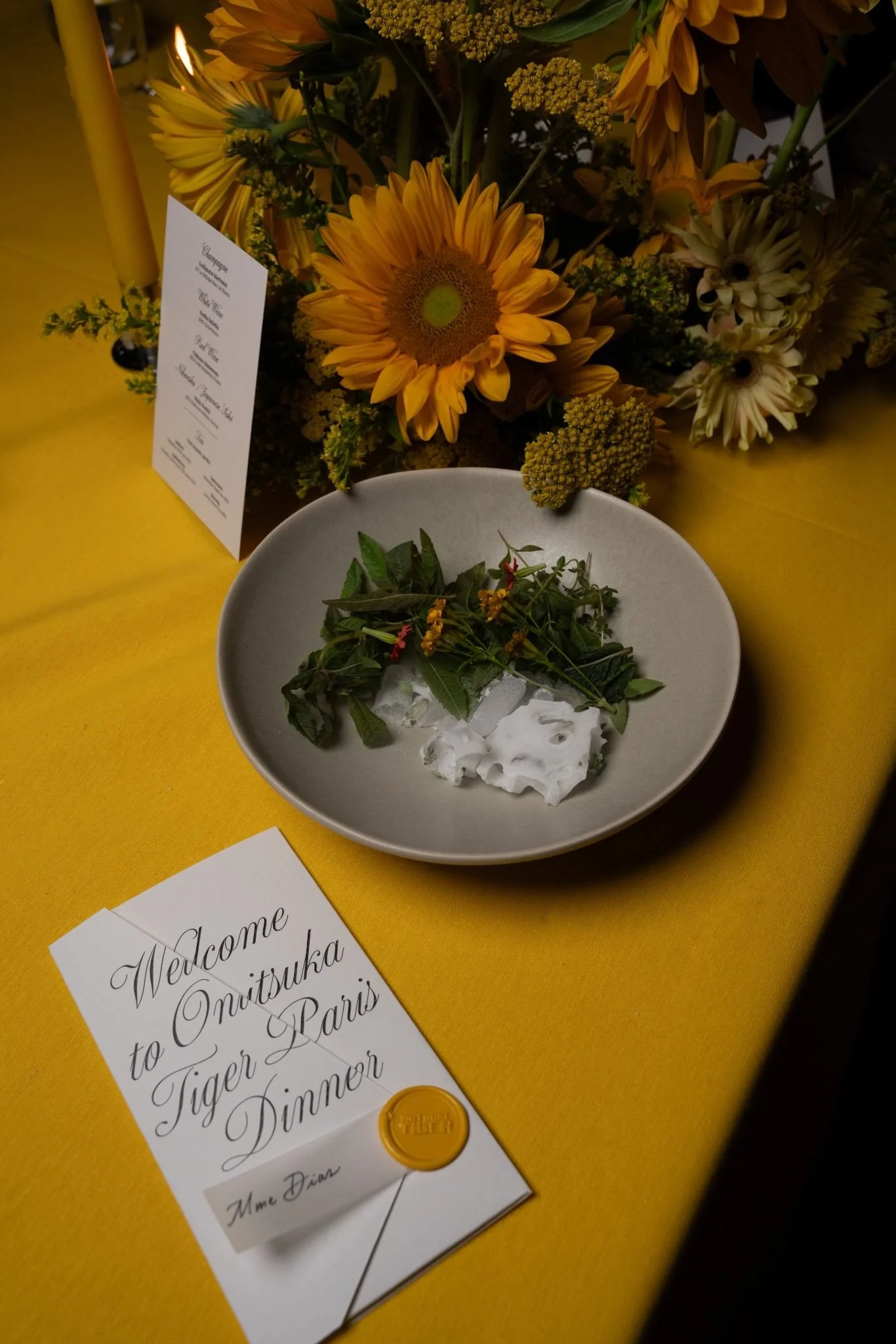 A table display featuring a bouquet of yellow and white flowers and a white card with cursive writing. The card includes a welcome message for a dinner event in Paris. A small white dish contains green herbs and white foam, adorned with a sprig of ye