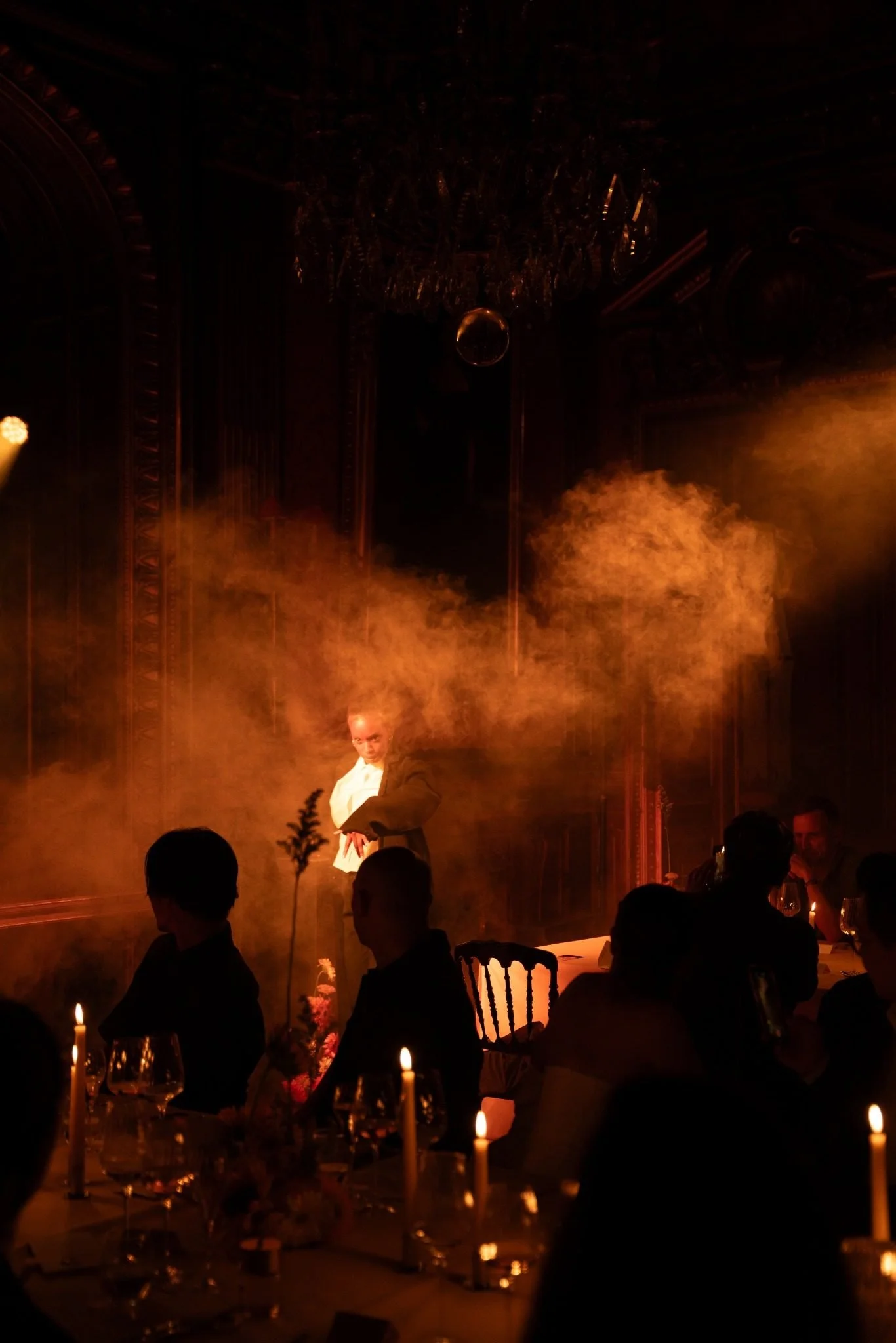 A performer in formal attire performs a dance on a dimly lit stage decorated with candles and floral arrangements, with an ornate ceiling and chandelier overhead, in an elegant ballroom setting.