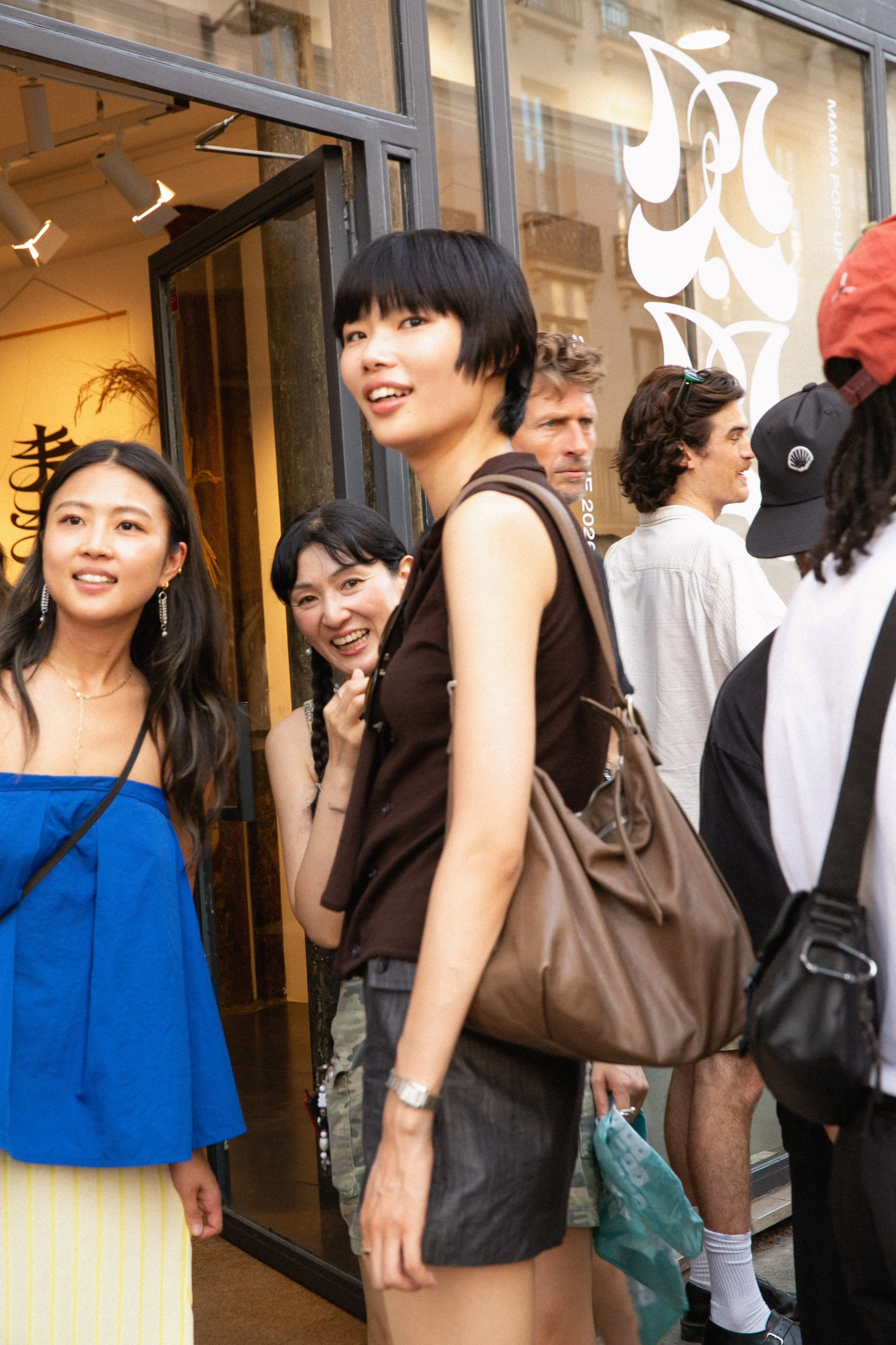Line of people standing outside a store entrance, some with bags, some engaged in conversation, and a woman smiling in the foreground wearing a sleeveless dark top and shorts. The storefront has glass windows with white decorative designs.