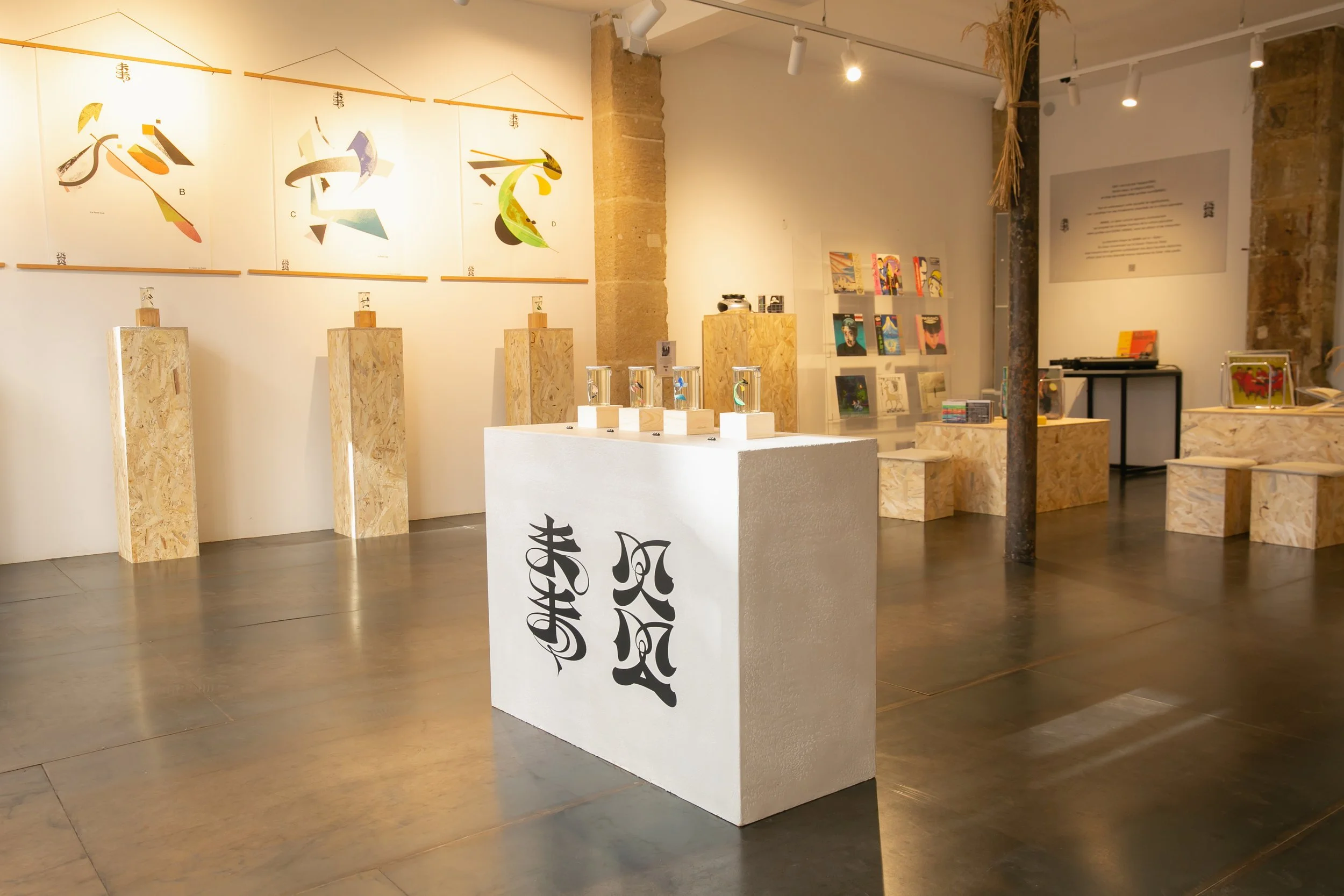 An art gallery with colorful abstract paintings on the wall, a white display table with small glass sculptures, and books on a bookshelf. The space features exposed brick columns and wooden pedestal stands.