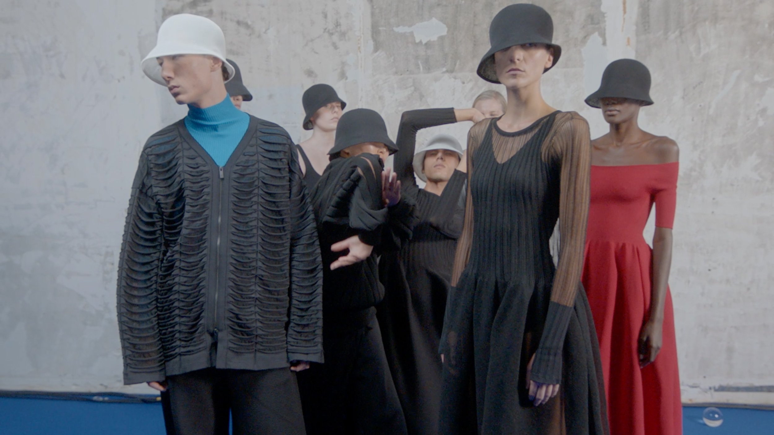 Six models stand against a gray, textured wall, wearing modern, avant-garde fashion with wide-brimmed hats in black, white, and gray, and dresses in black, red, and striped patterns.