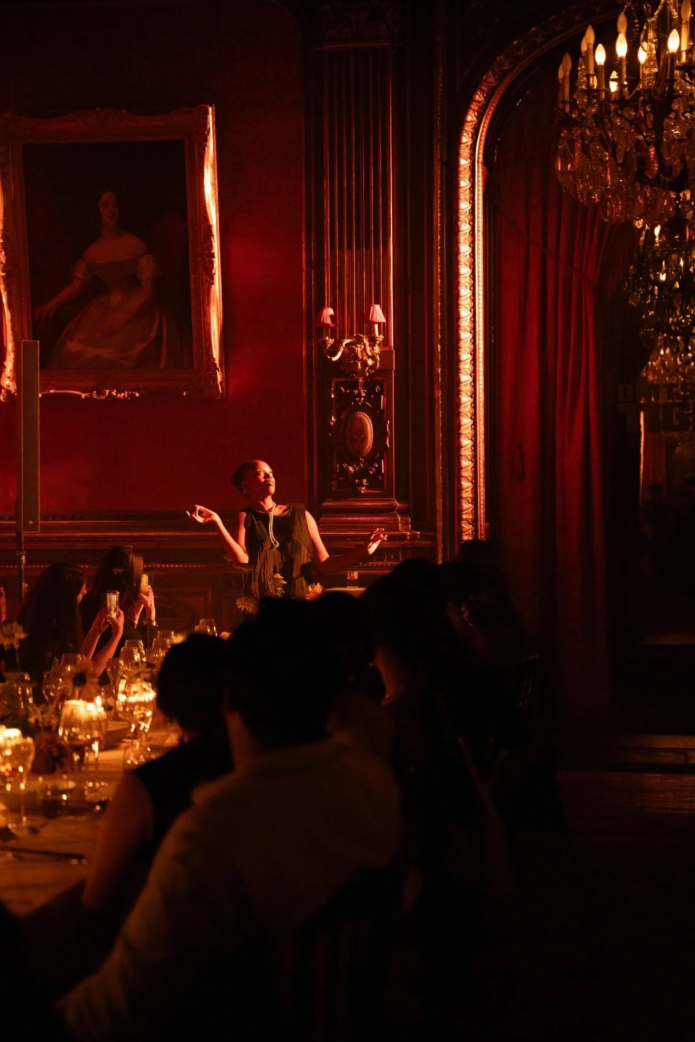 An elegant woman speaking at a formal dinner in a richly decorated room with red curtains, a chandelier, and a large framed portrait on the wall.