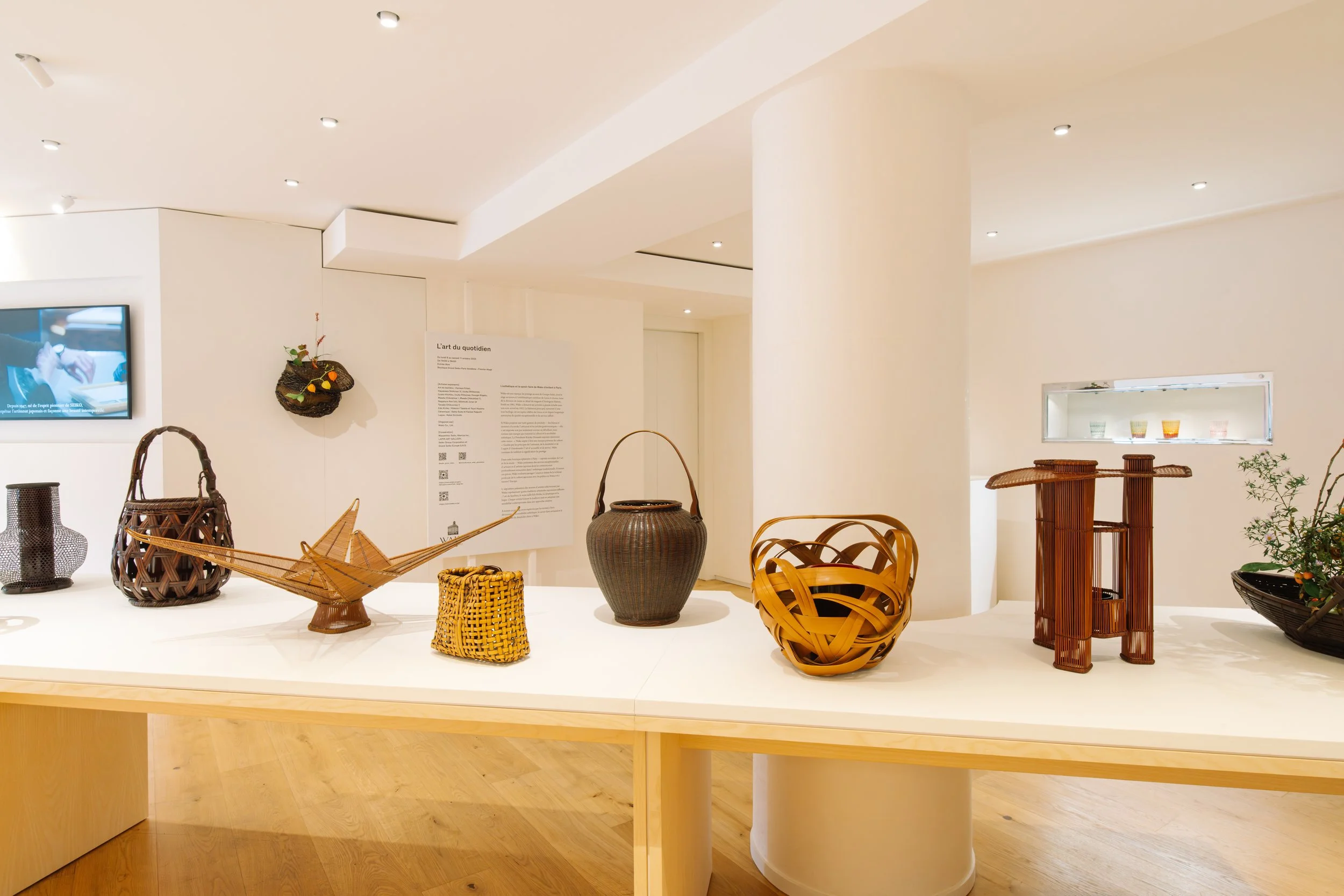 A display of woven baskets and decorative pottery on a white table in a bright art gallery with white walls and wooden floors.