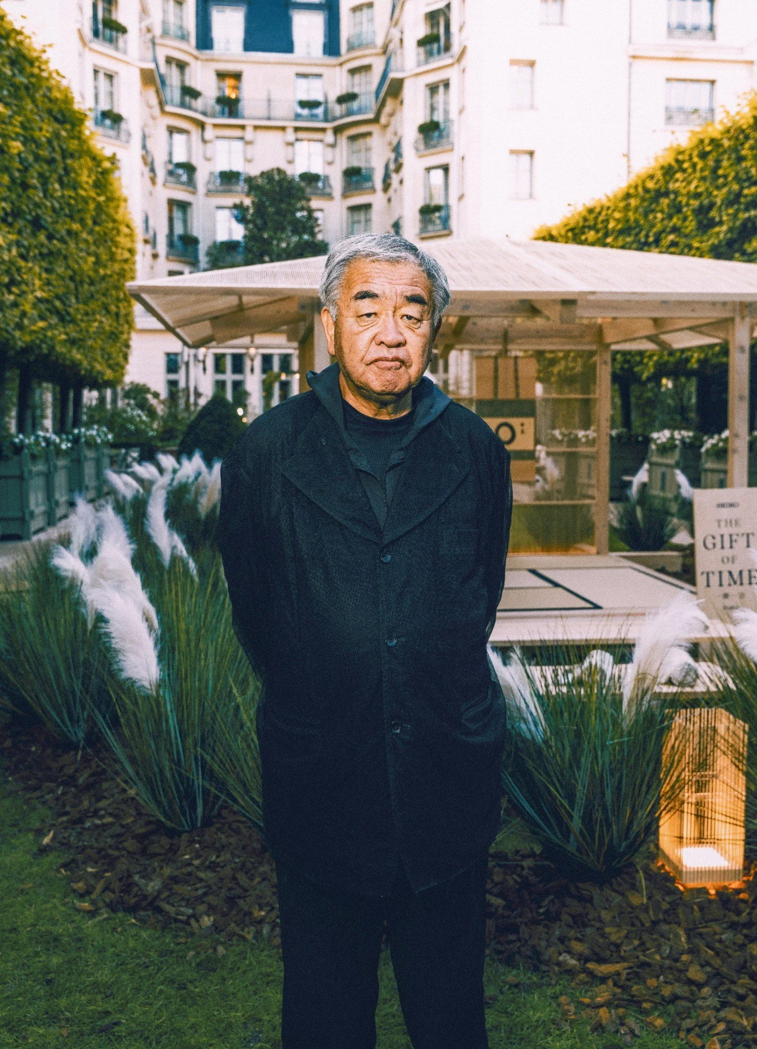 A man with gray hair standing outdoors in a garden area with a modern apartment building in the background, with plants and a wooden structure nearby.