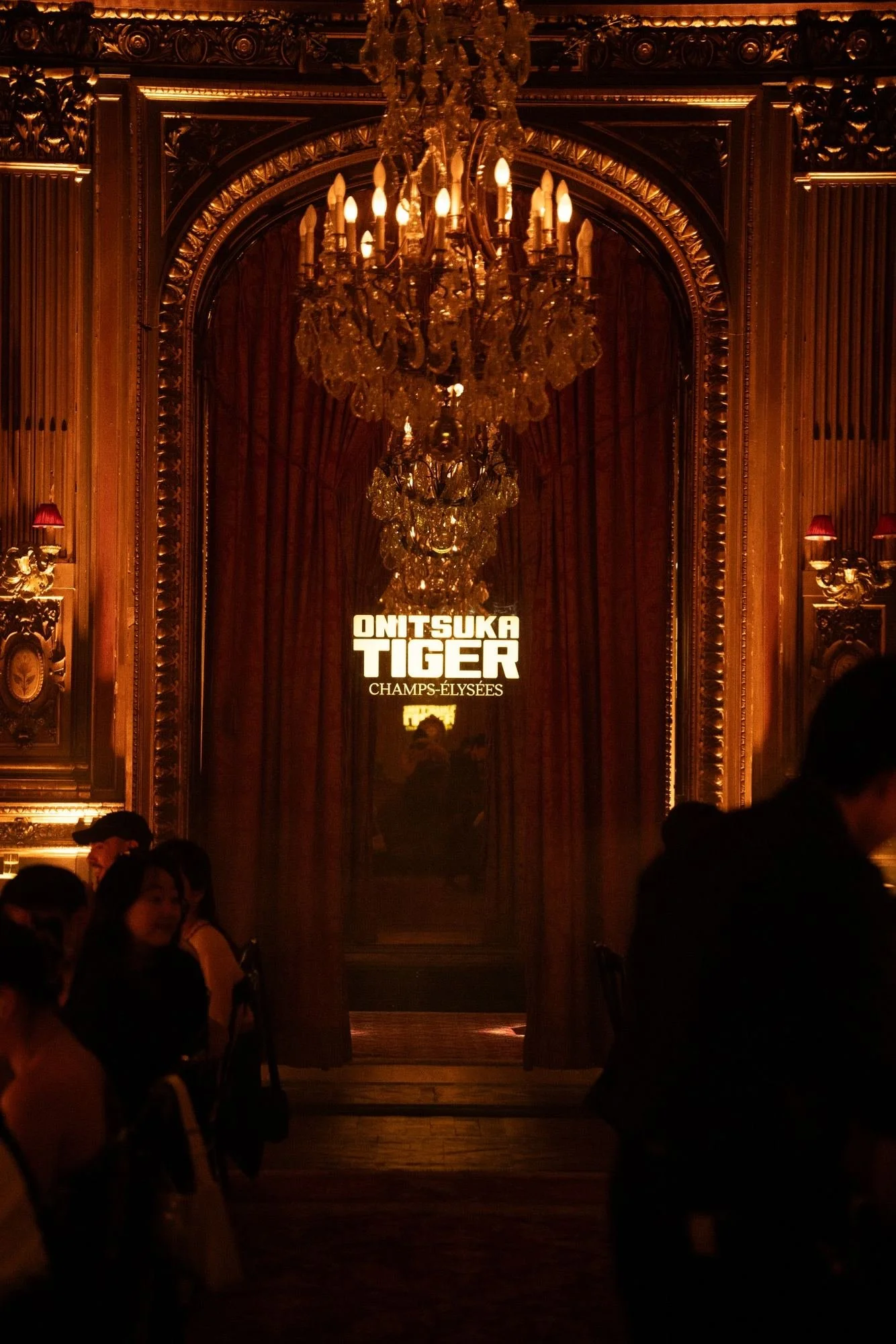 An ornate ballroom with a large chandelier, red curtains, and a sign that reads "Omi Tsuka Tiger Champs-Élysées." Several people are seen in the darker foreground, some appearing to be waiting or talking.