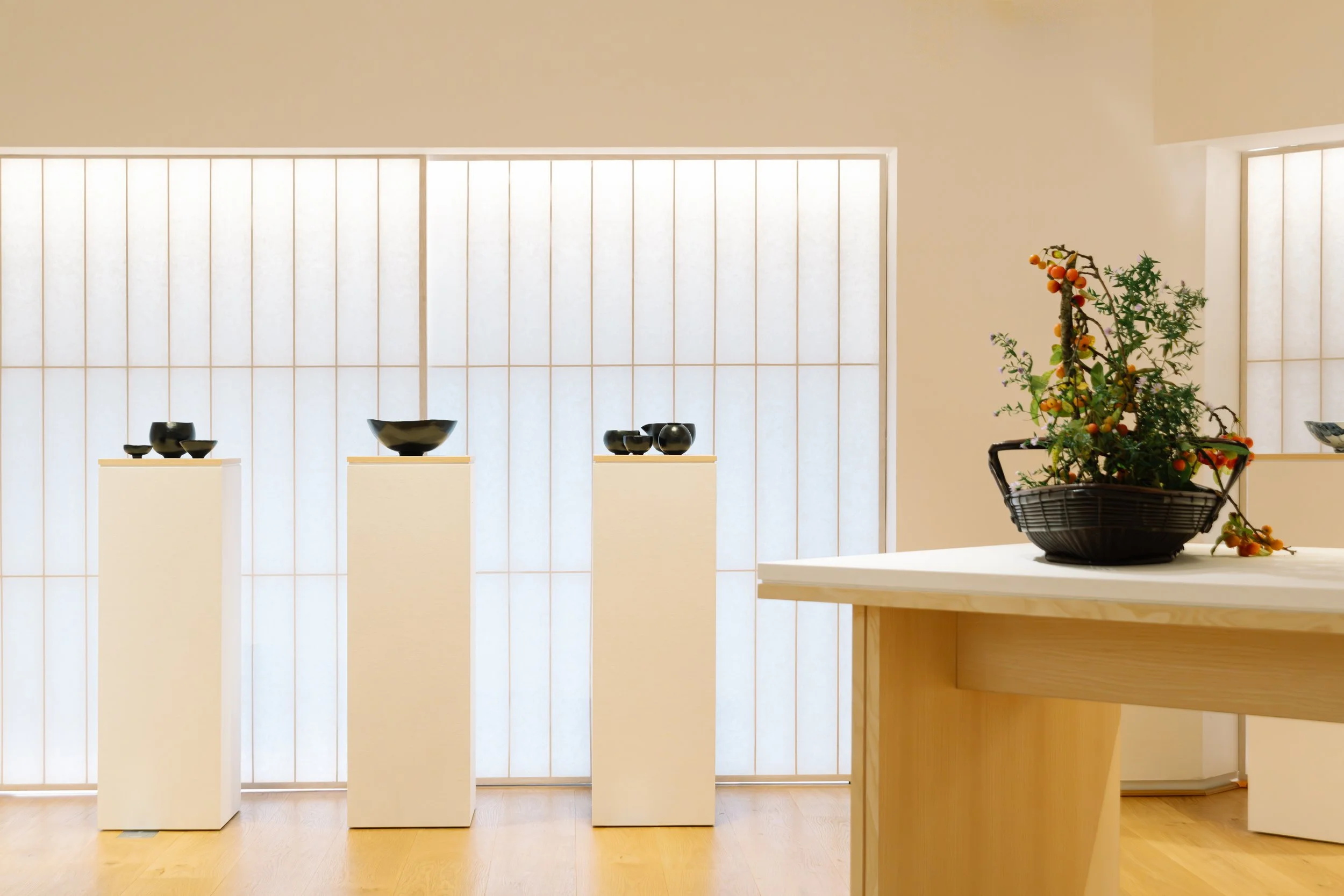 A display of black ceramic bowls on white pedestals against a shoji paper screen, with a bonsai tree with small orange fruit on a wooden table in the foreground.