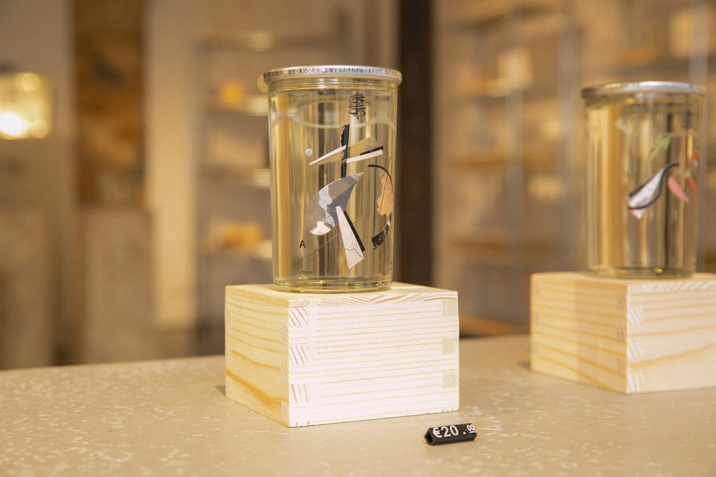 A glass of water with abstract and geometric artwork attached to its surface, displayed on a wooden block with a price tag of €20.