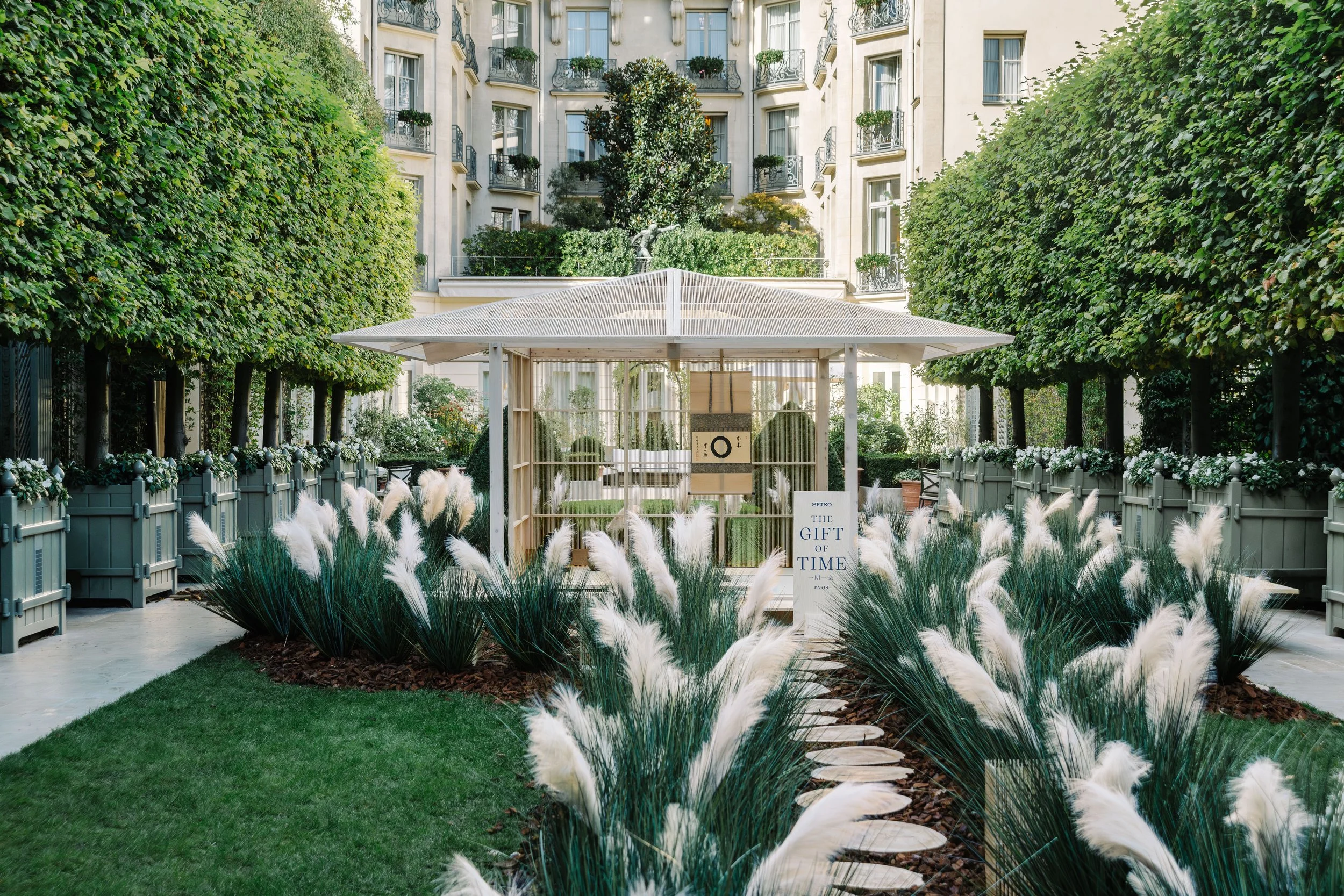 Ritz Paris courtyard with a tea house build by Kengo Kuma, lush green hedges, decorative white pampas grasses, and an elegant cream-colored apartment building with balconies and ornate railings in the background.