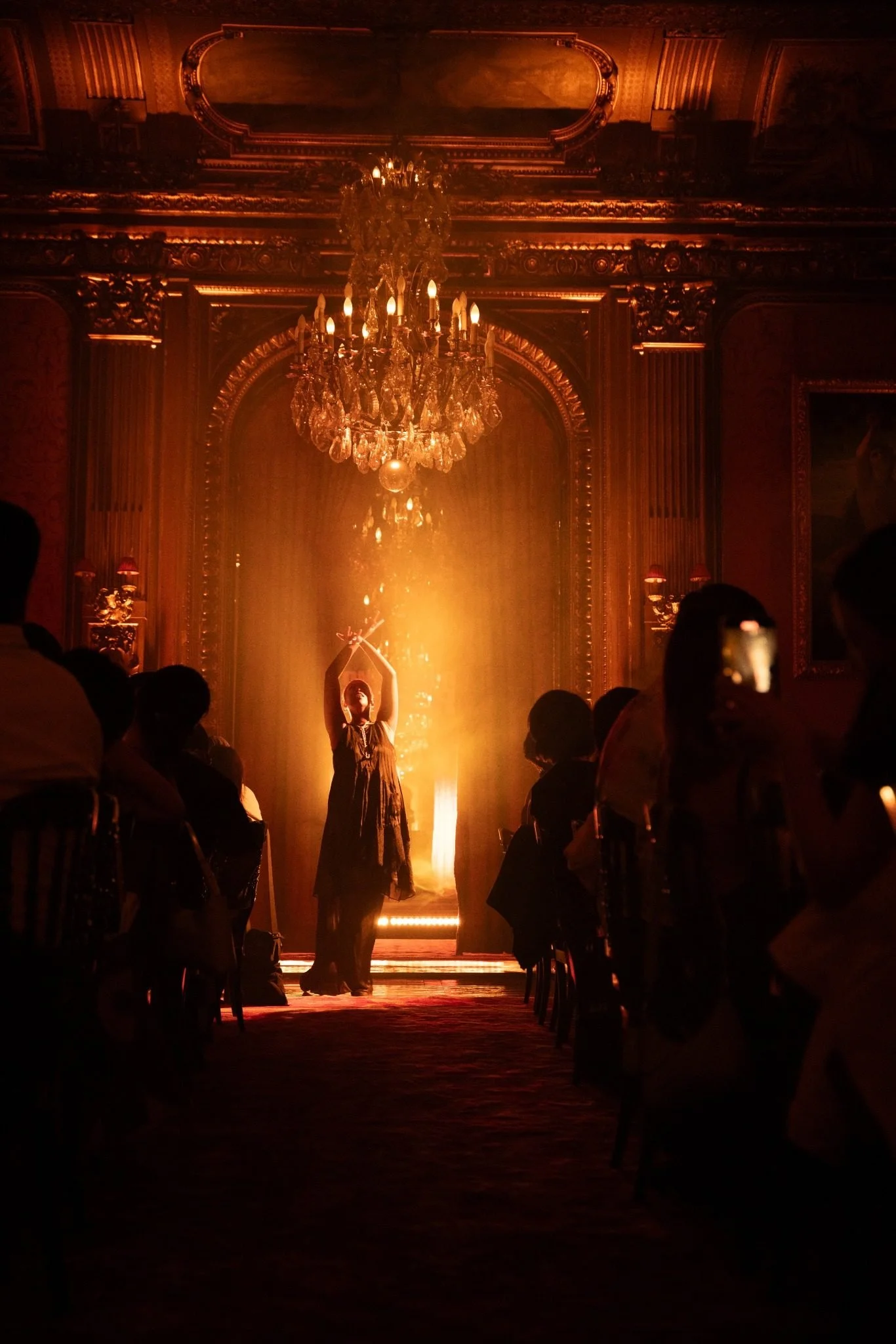 A woman in a black dress stands with arms raised in front of an ornate, chandelier-lit room, with an audience seated on either side, in front of a tall mirror and opulent gold decorations.