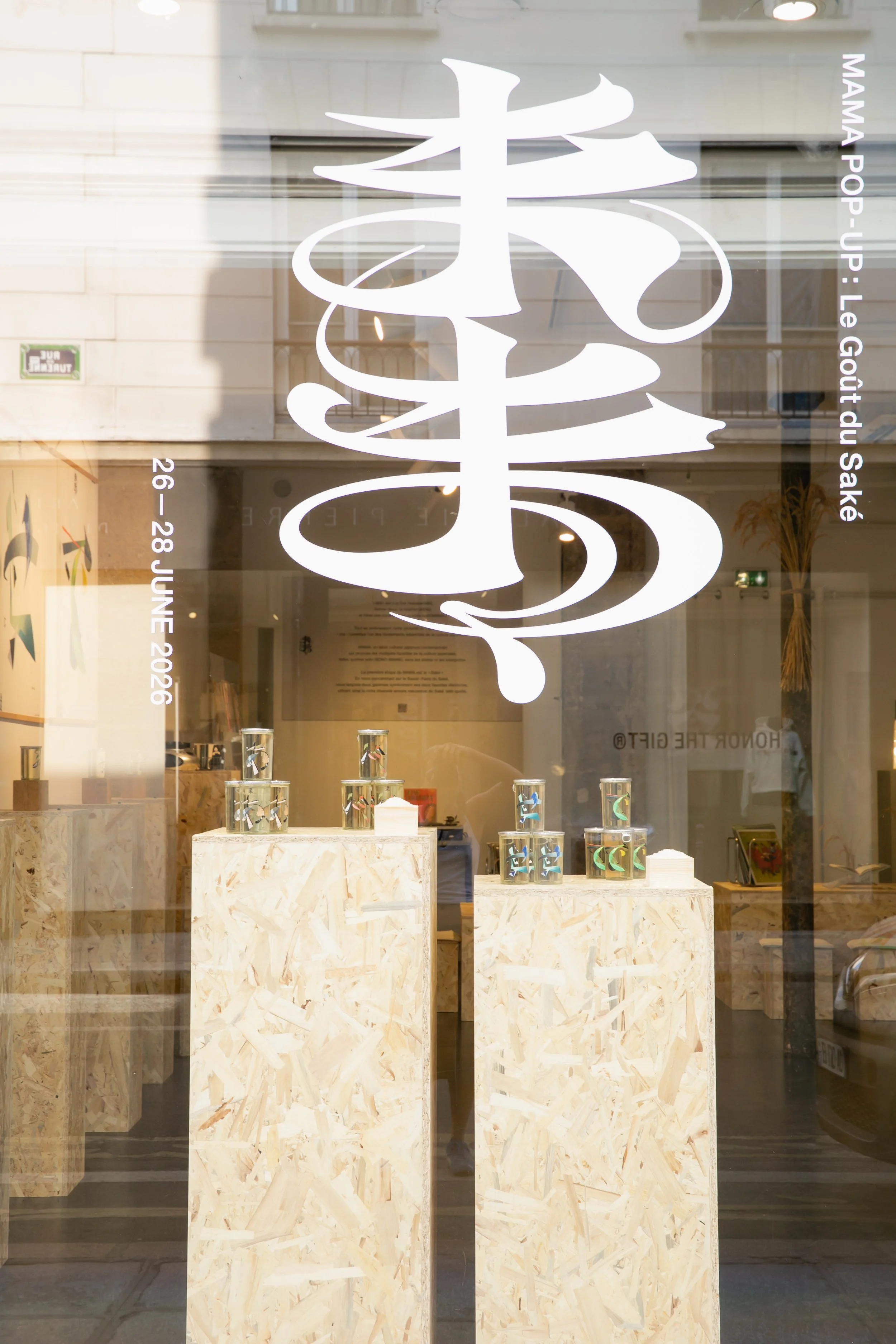 Store window display featuring glassware and decorative items on light wood pedestals, with a large stylized white logo and text in French indicating the store's name, location, and dates.