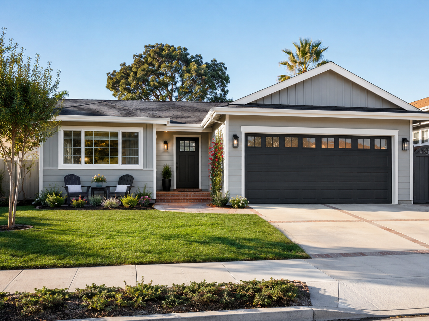 Modern house with gray exterior, black garage door, front porch with seating and plants, and well-maintained lawn under blue sky.