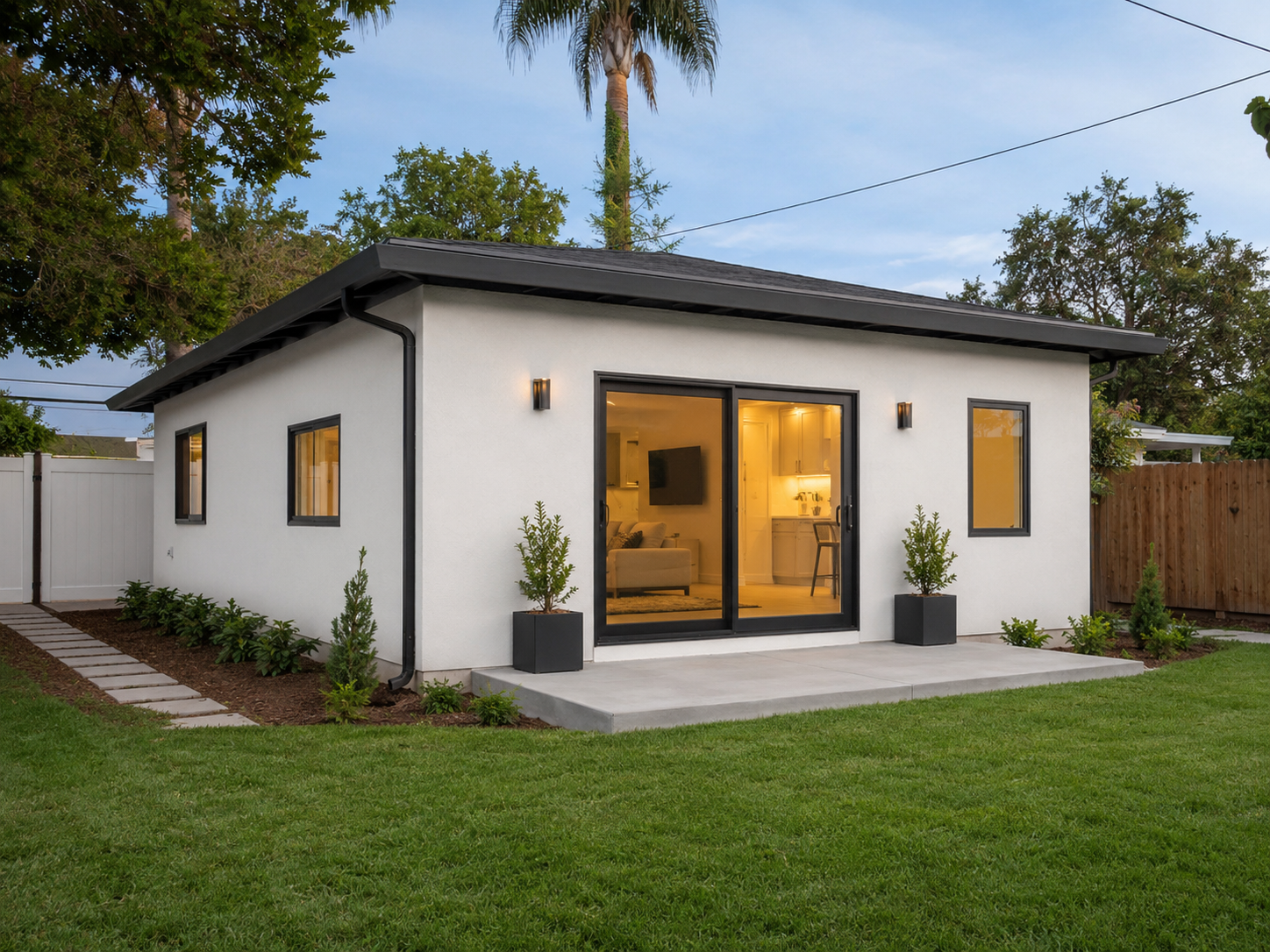 Modern small house with white exterior walls, large sliding glass door, and black window frames. Green lawn and landscaped garden with small trees in black planters outside, evening lighting inside, wooden fence in background, tall trees, and a cloudy sky.