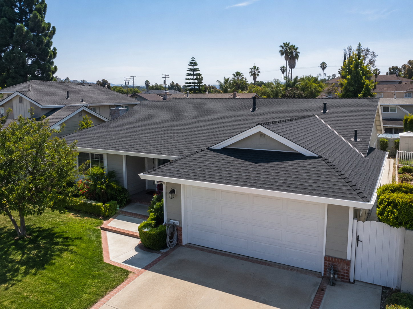 Aerial view of a suburban house with a gray shingle roof, white garage door, and surrounding trees and landscaping.