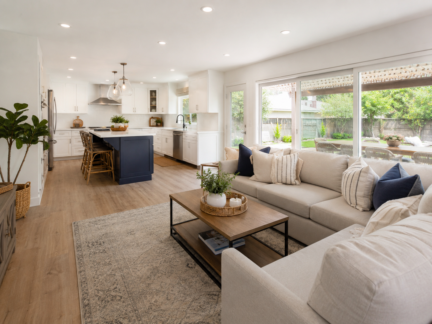 Open-concept living room and kitchen with white cabinetry, navy island, neutral sofa, large windows overlooking backyard with patio and green plants, wooden coffee table, area rug, and indoor plants.