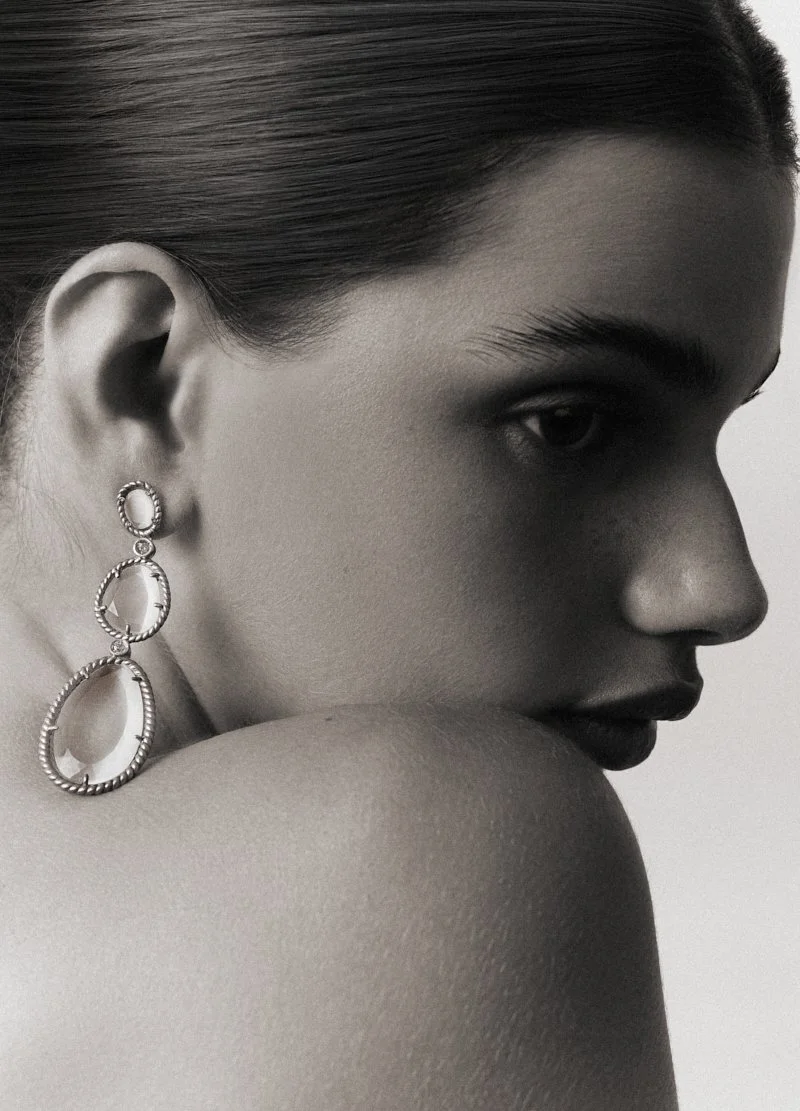 Portrait en noir et blanc d'une femme avec une tête légèrement tournée, portant une boucle d'oreille pendante ornée de cercles et de détails texturés.