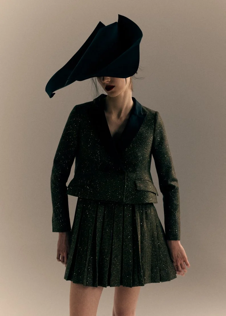 Femme portant un costume noir scintillant avec une robe plissée et un grand chapeau noir.