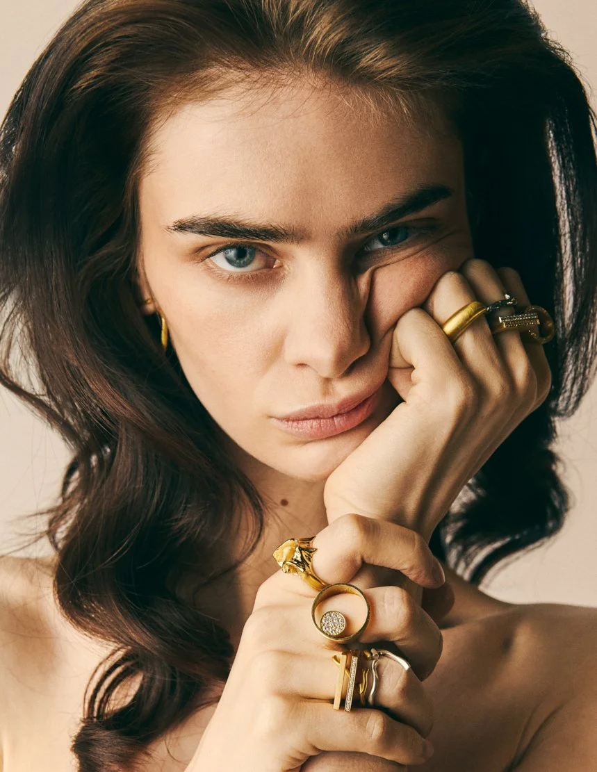 Portrait d'une femme aux cheveux bruns, yeux bleus, portant plusieurs bijoux en or, posant avec la main près du visage.