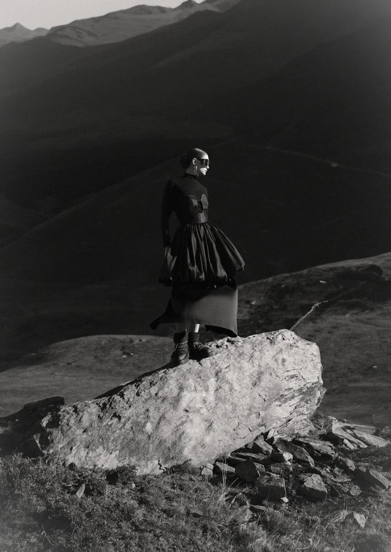 Une femme habillée en noir, portant un vêtement ample avec une jupe, se tient sur un gros rocher dans un paysage montagneux, en noir et blanc.