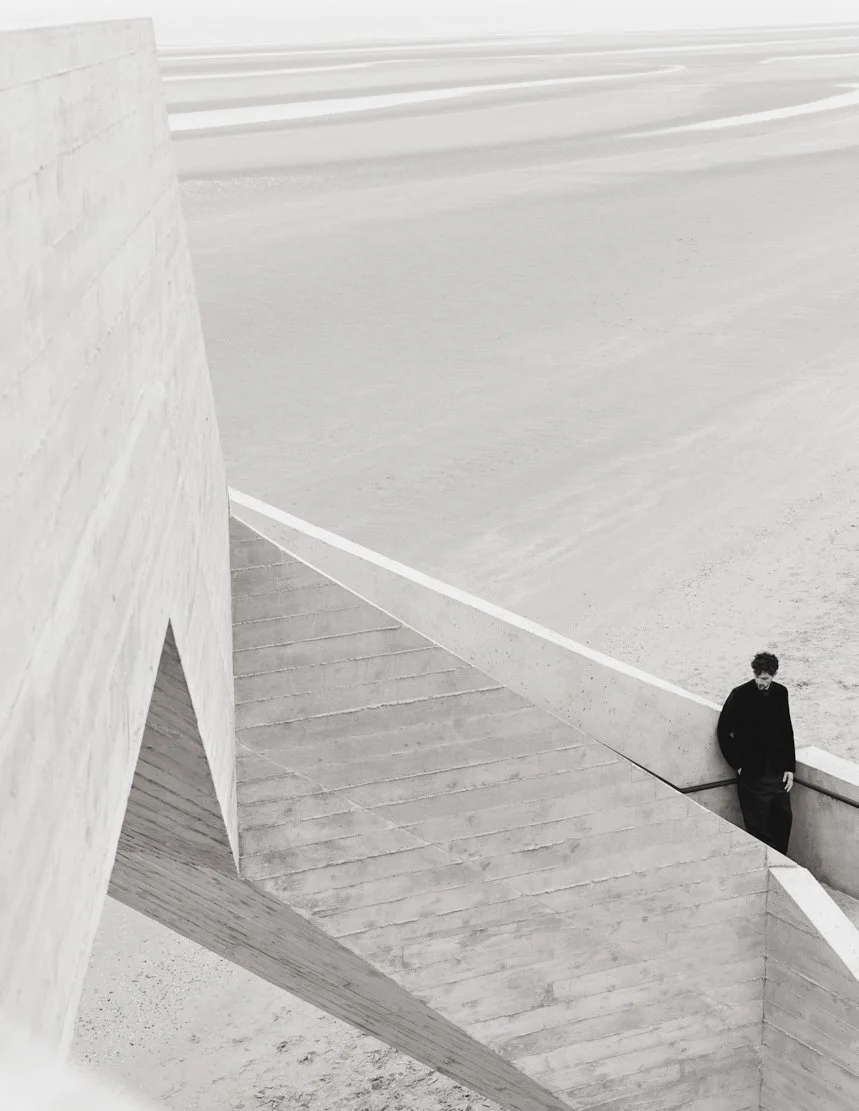 Un homme habillé en noir se tient contre un mur dans une architecture moderne, avec des structures en béton et un espace ouvert en arrière-plan.