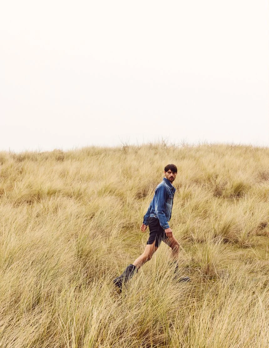 Homme marchant dans une prairie d'herbes hautes, portant un jean et des bottes, regardant vers l'objectif.