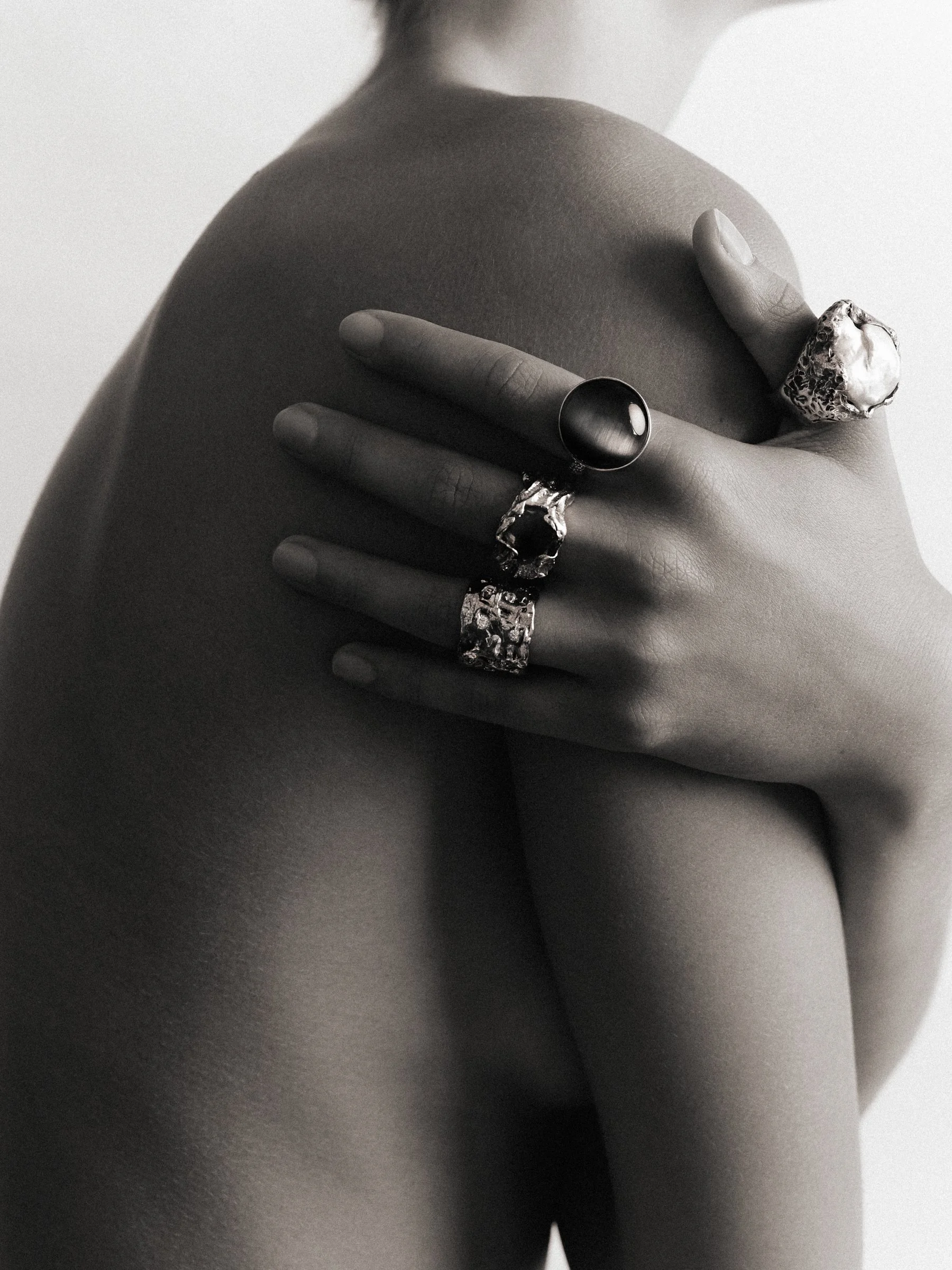A close-up black and white photo of a woman's hand with rings, touching her shoulder and back.