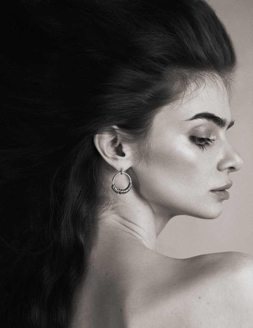 portrait en noir et blanc d'une femme avec un grand volume de cheveux foncés, portant un boucle d'oreille en cercle, vue en profil avec regard baissé.