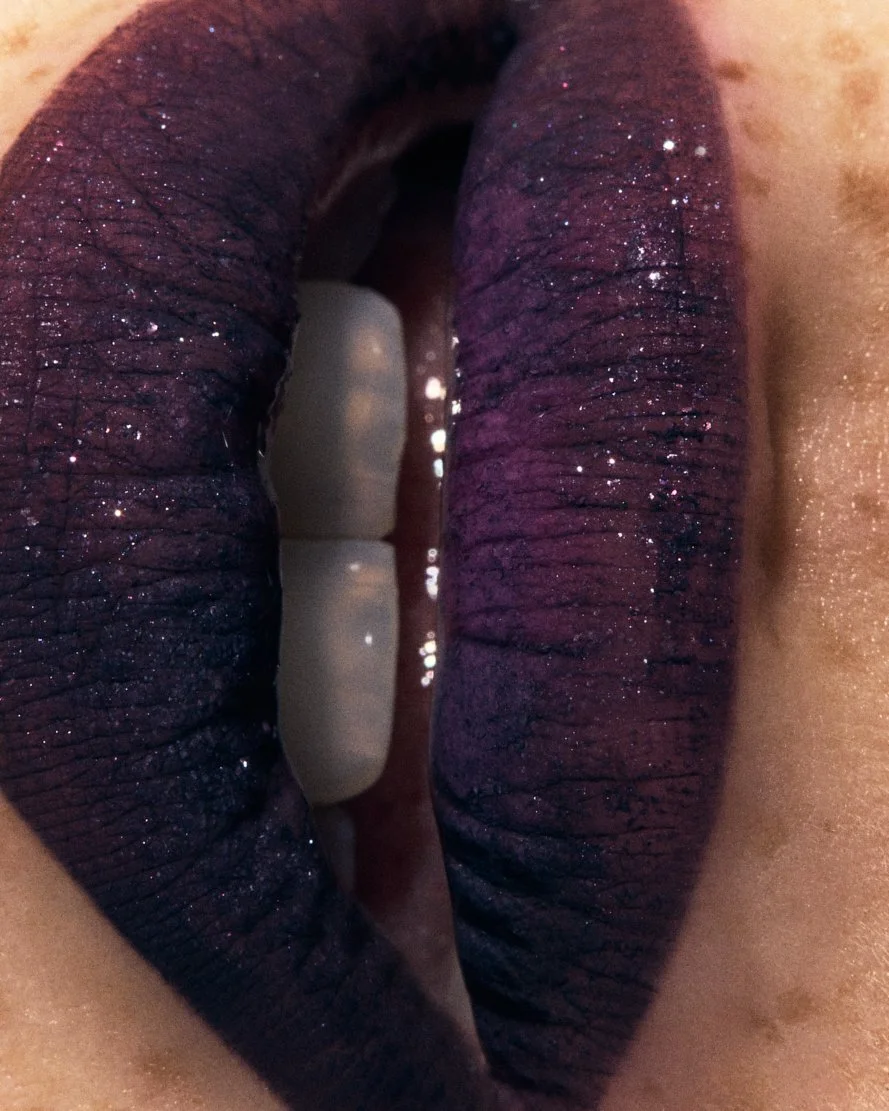 Lèvres pulpeuses avec maquillage violet foncé et paillettes, légèrement entrouvertes, révélant des dents visibles.