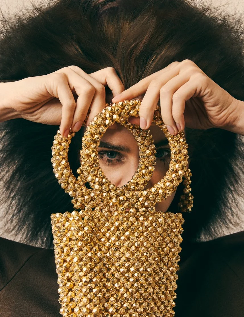 Une personne avec des cheveux volumineux tient des accessoires dorés en forme de cercles devant son visage, créant un effet artistique.