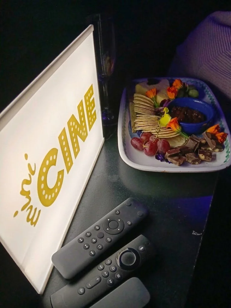 Party platter with crackers, grapes, chocolates, and a dip, next to a 'MINI CINE' sign, with remote controls on a dark table.