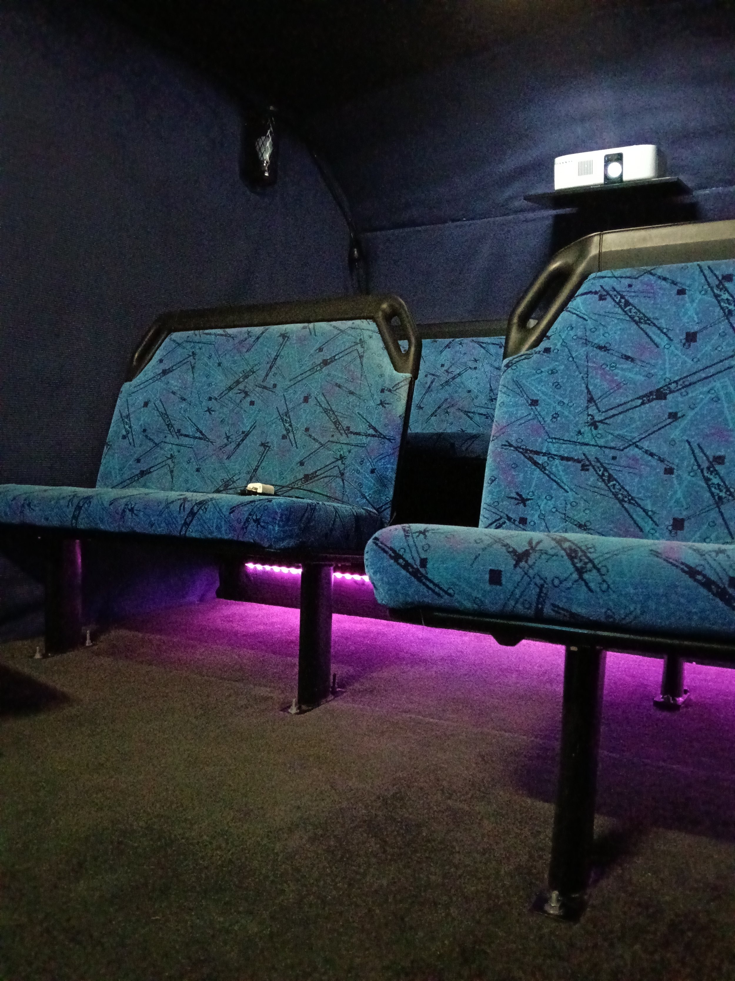 Empty cinema seats with blue patterned fabric, illuminated by purple accent lighting underneath, with black headrests and a dark background.