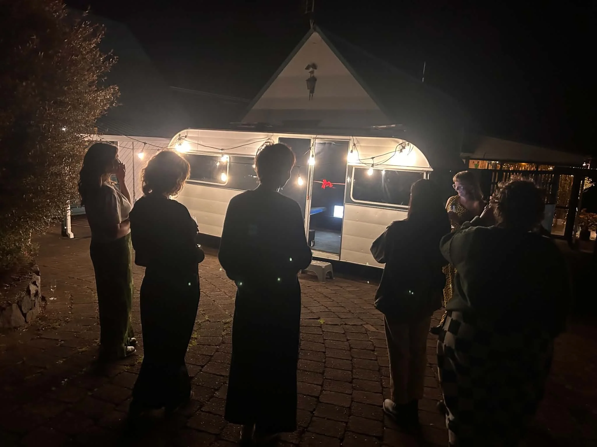 Six people are gathered outdoors at night, standing in front of a small trailer with string lights hanging around its entrance. One person is taking a photo, while others are engaged in conversation or looking at the trailer.